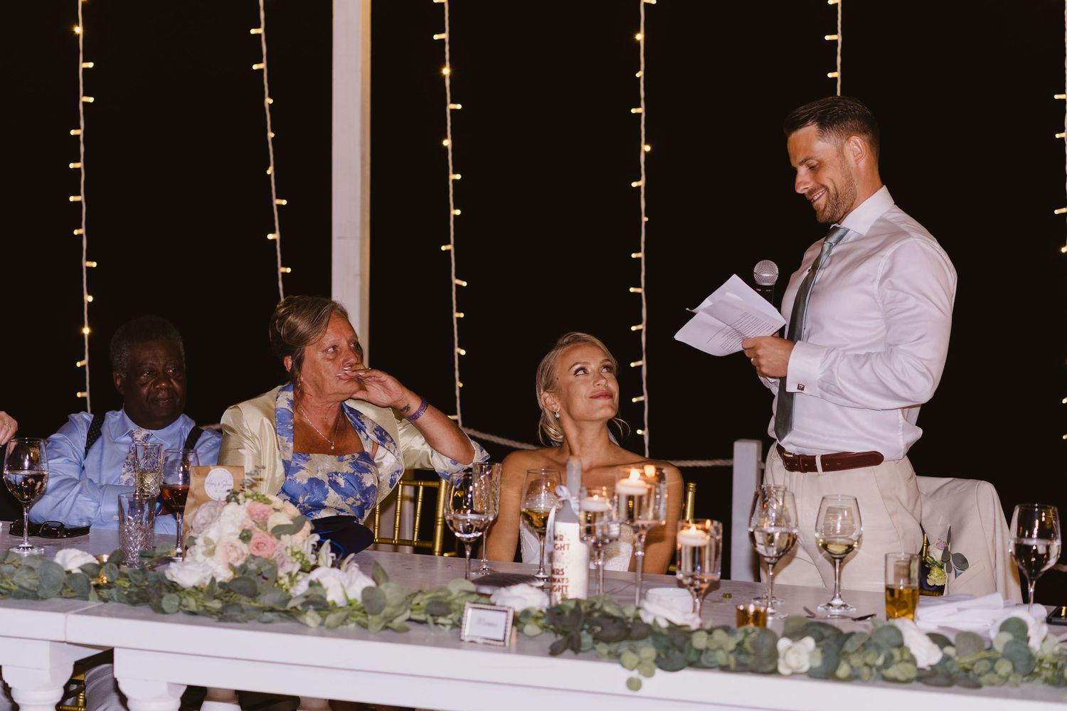 Wedding reception speech elicits laughter from guests at head table.