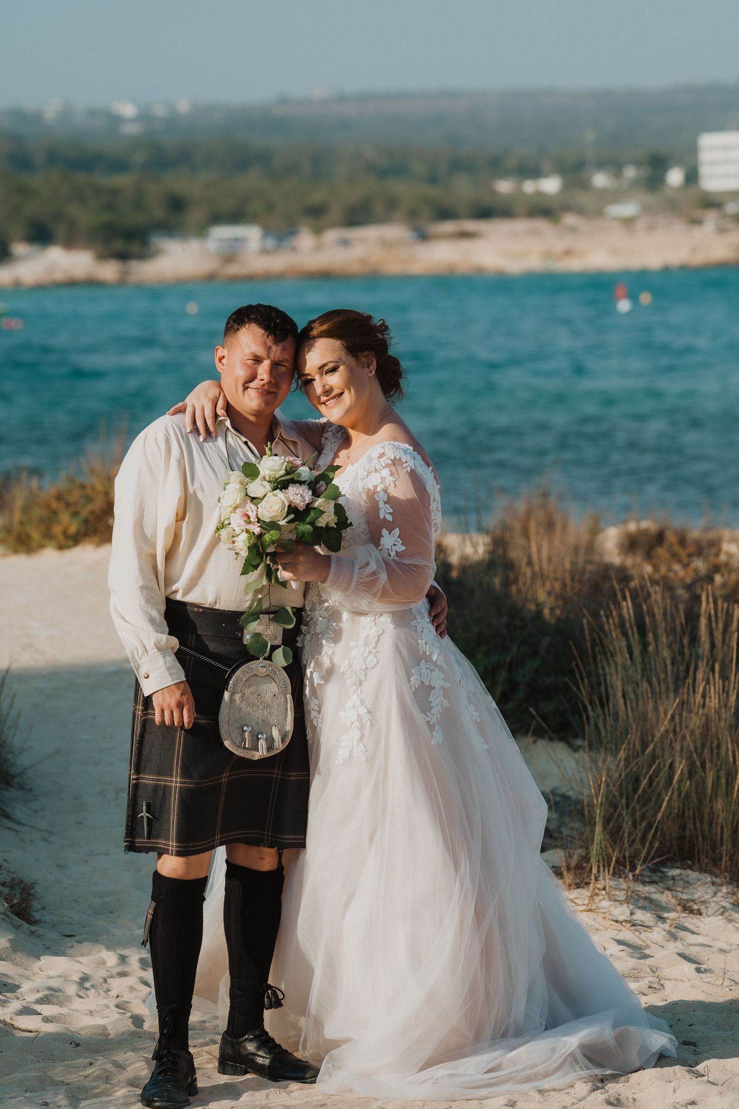 A couple in wedding attire embraces on a sandy beach with turquoise waters in the background.