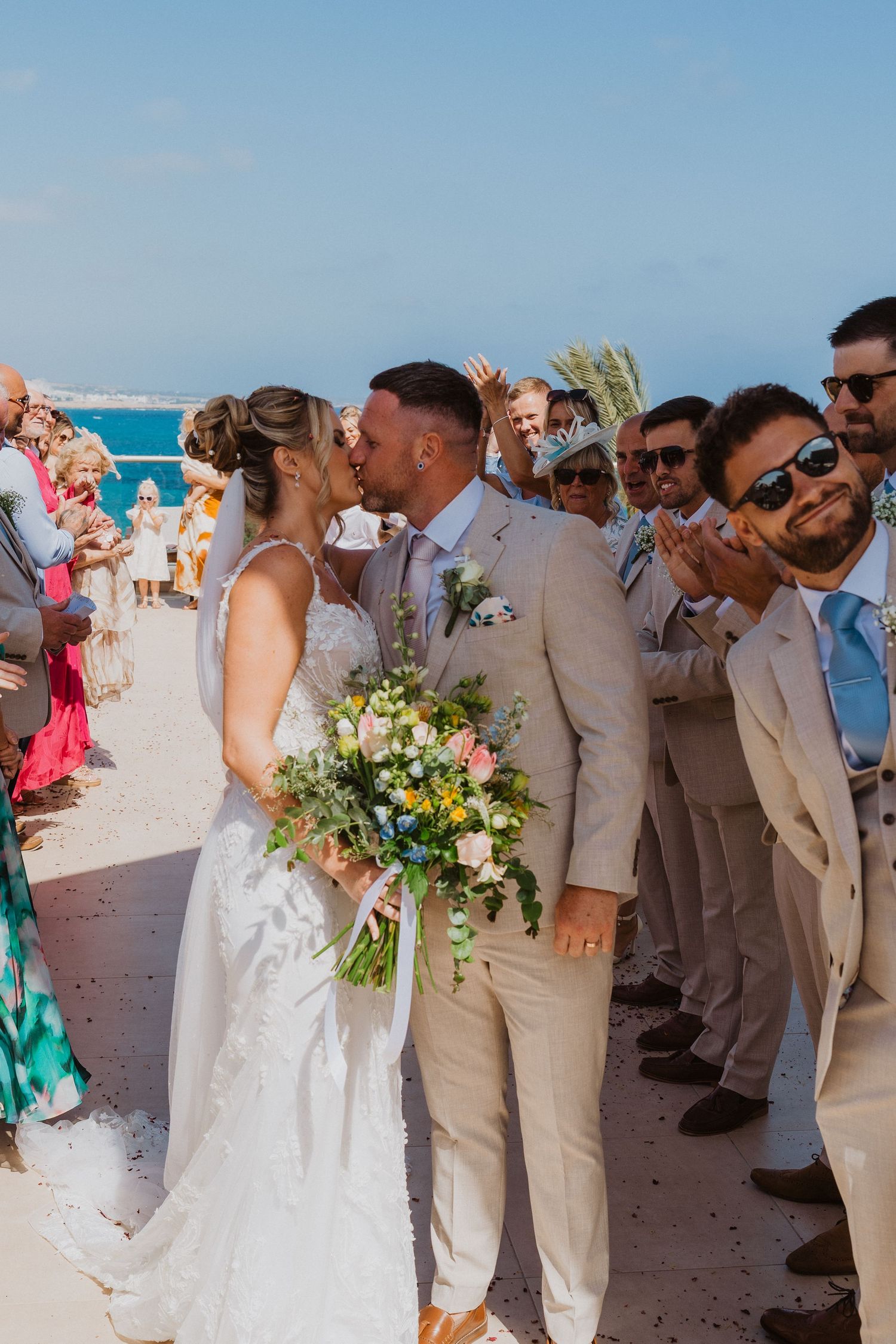 Wedding party in light beige suits celebrates on a sunny beach with blue ocean views and tropical flowers.