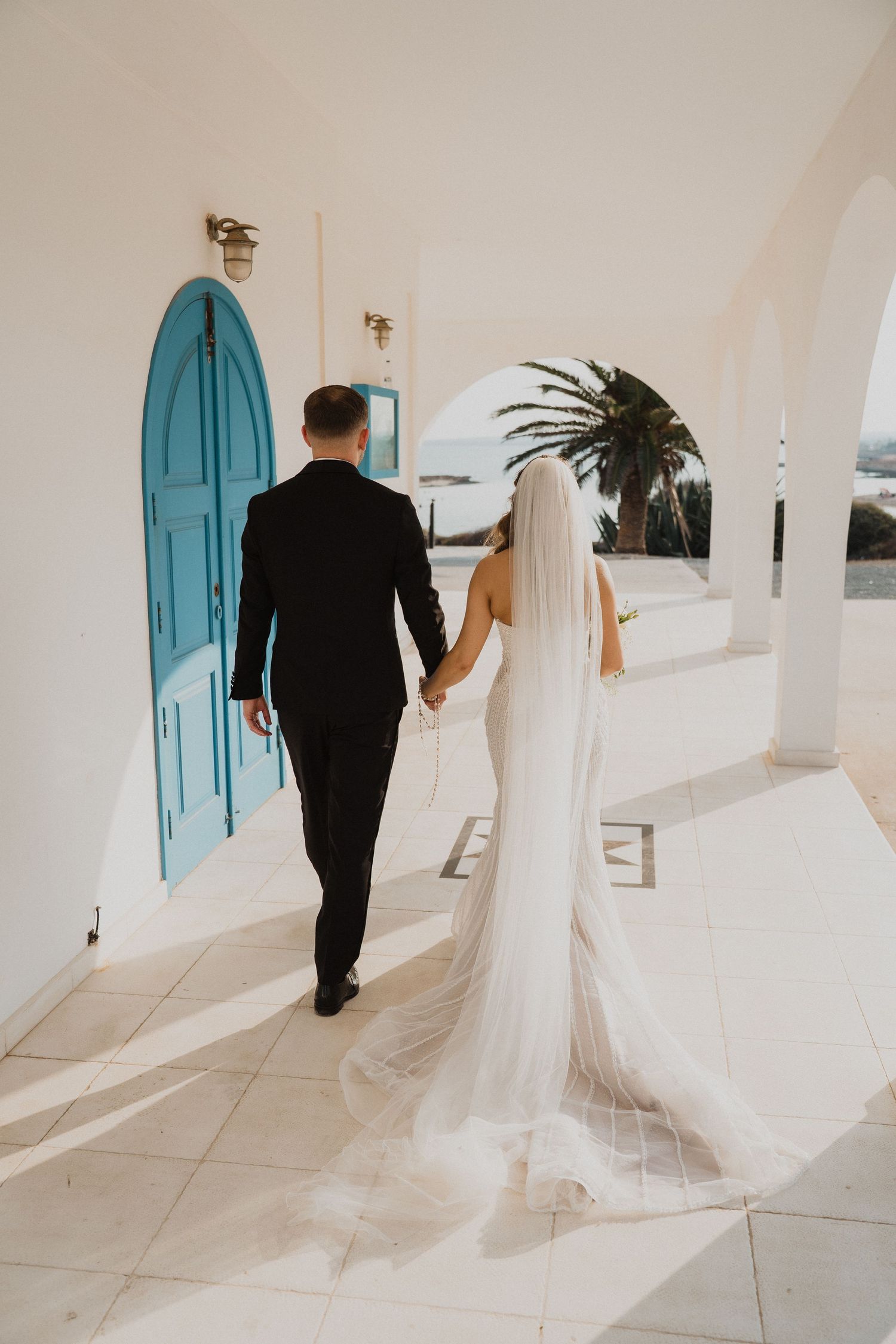 Bride and groom walking hand in hand along a white Mediterranean venue with turquoise doors and ocean views.