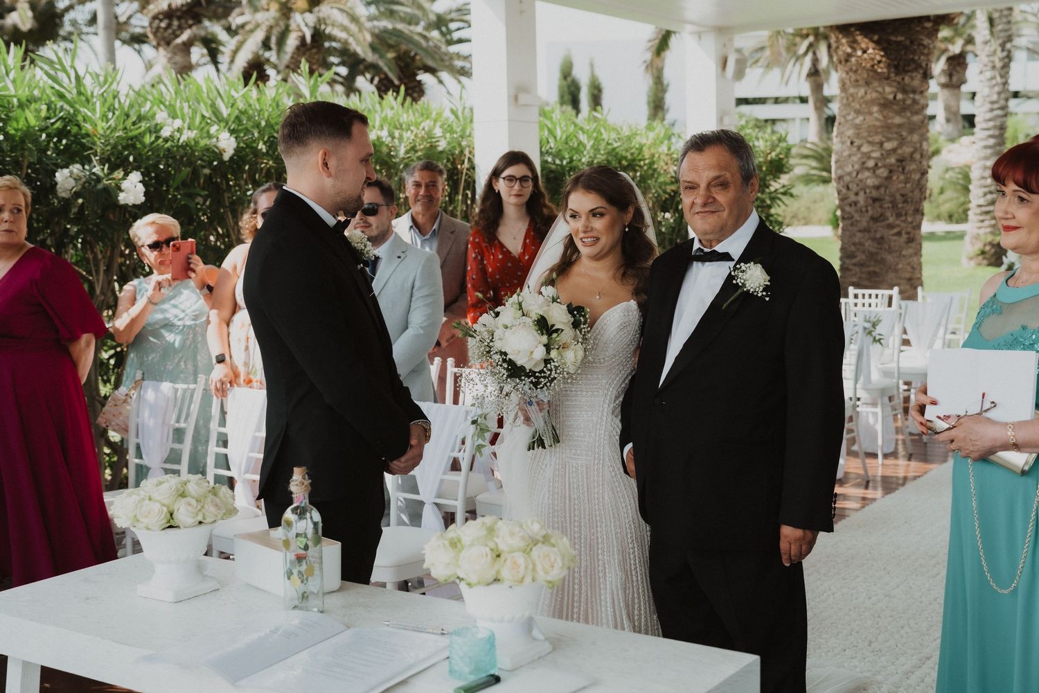 Bride in beaded gown with bouquet stands with groom and father at outdoor wedding ceremony.