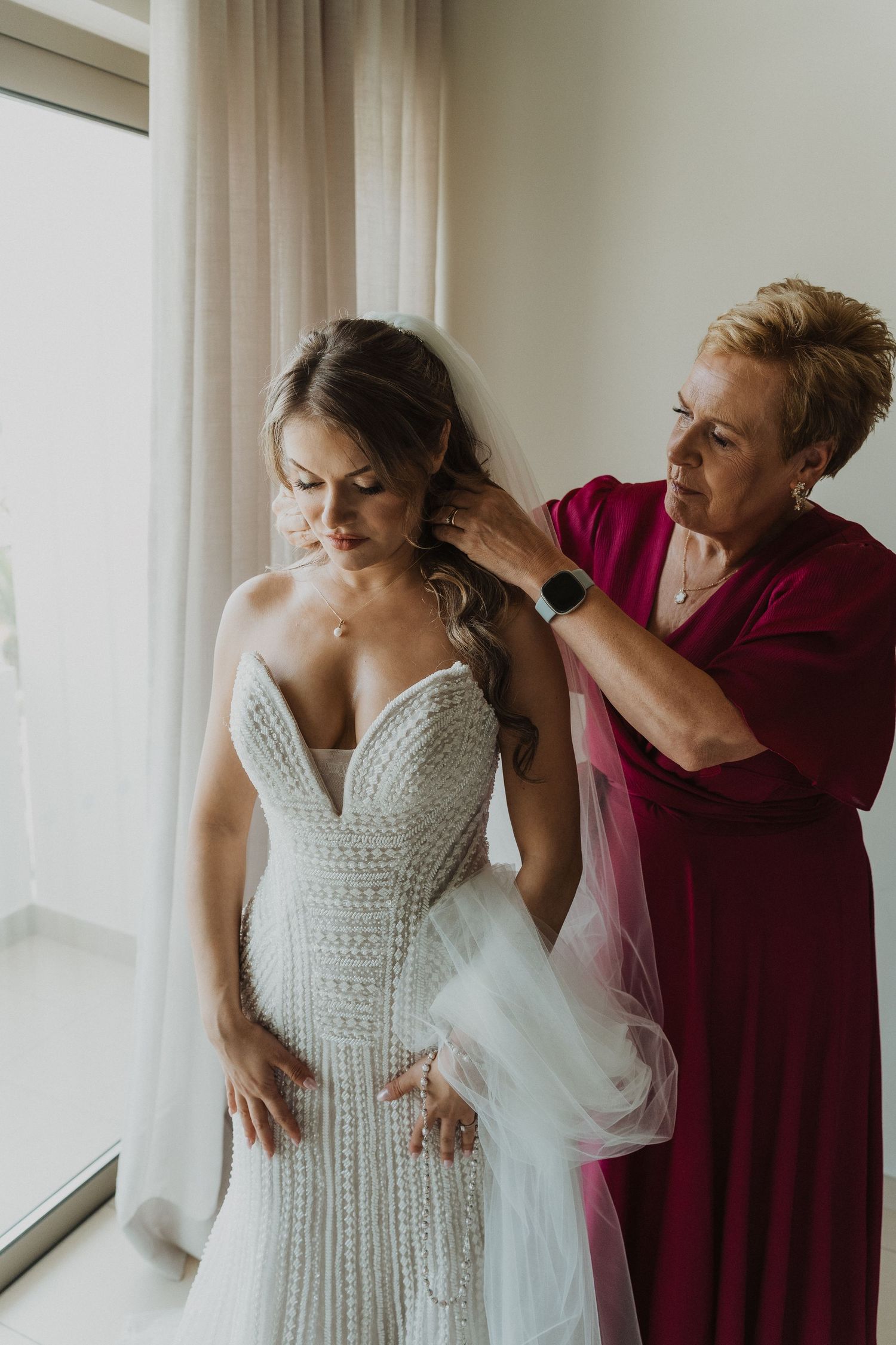 A bride in a beaded mermaid wedding gown having her veil adjusted by an older woman in a red dress.