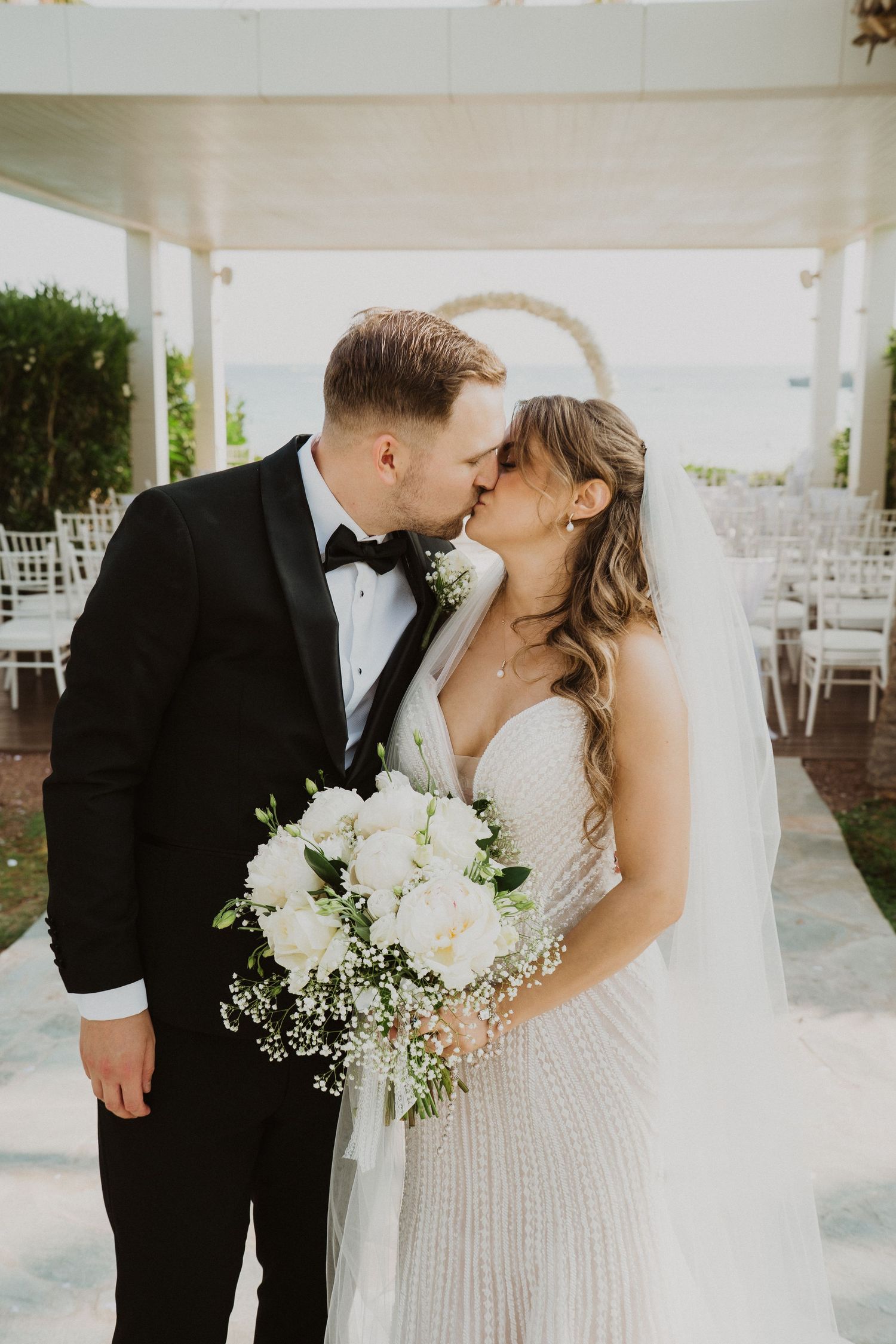 Bride and groom share a kiss at outdoor wedding ceremony with white chairs and ocean backdrop.