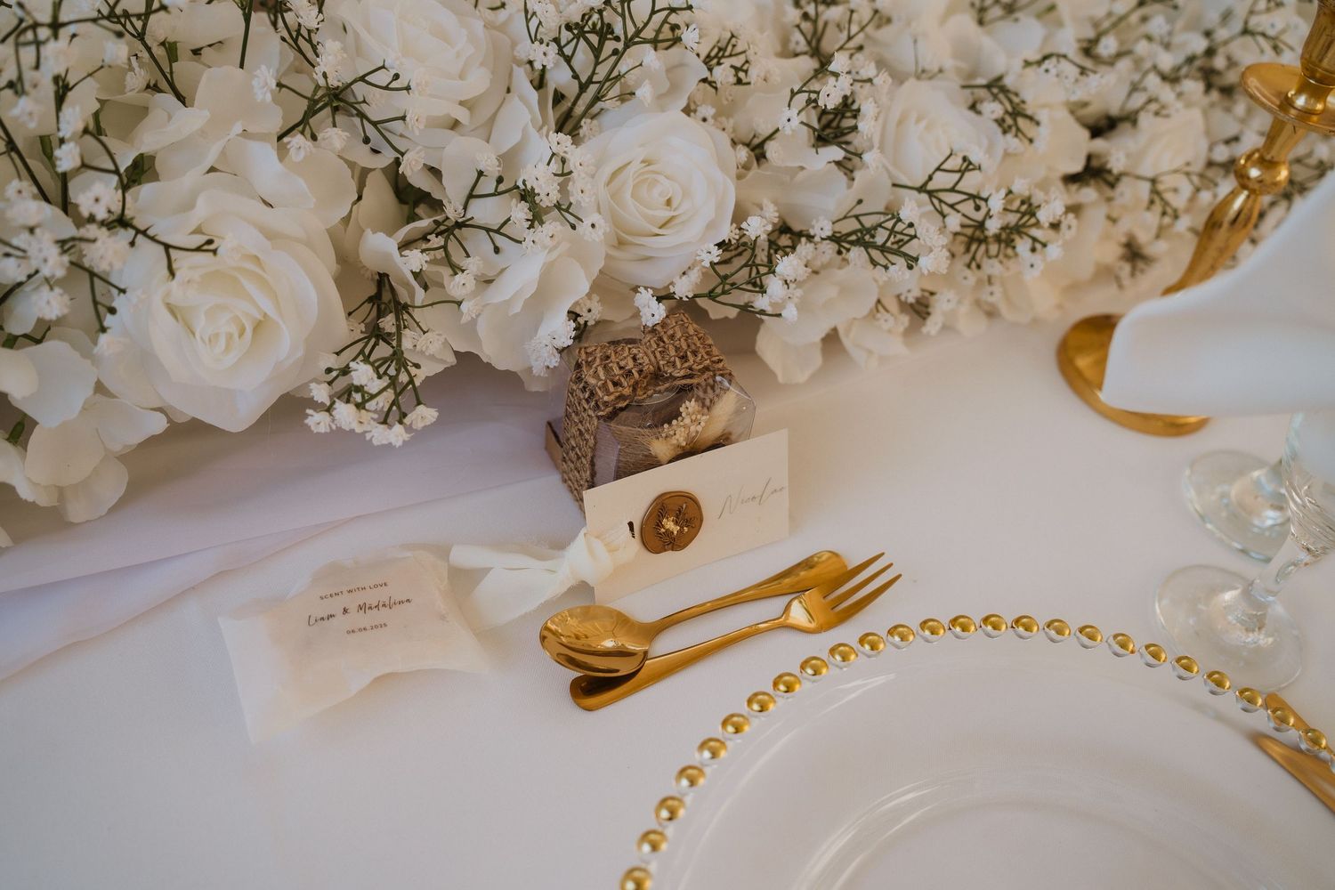 Elegant wedding table setting with white roses, gold cutlery, beaded charger plates and small gift favors.
