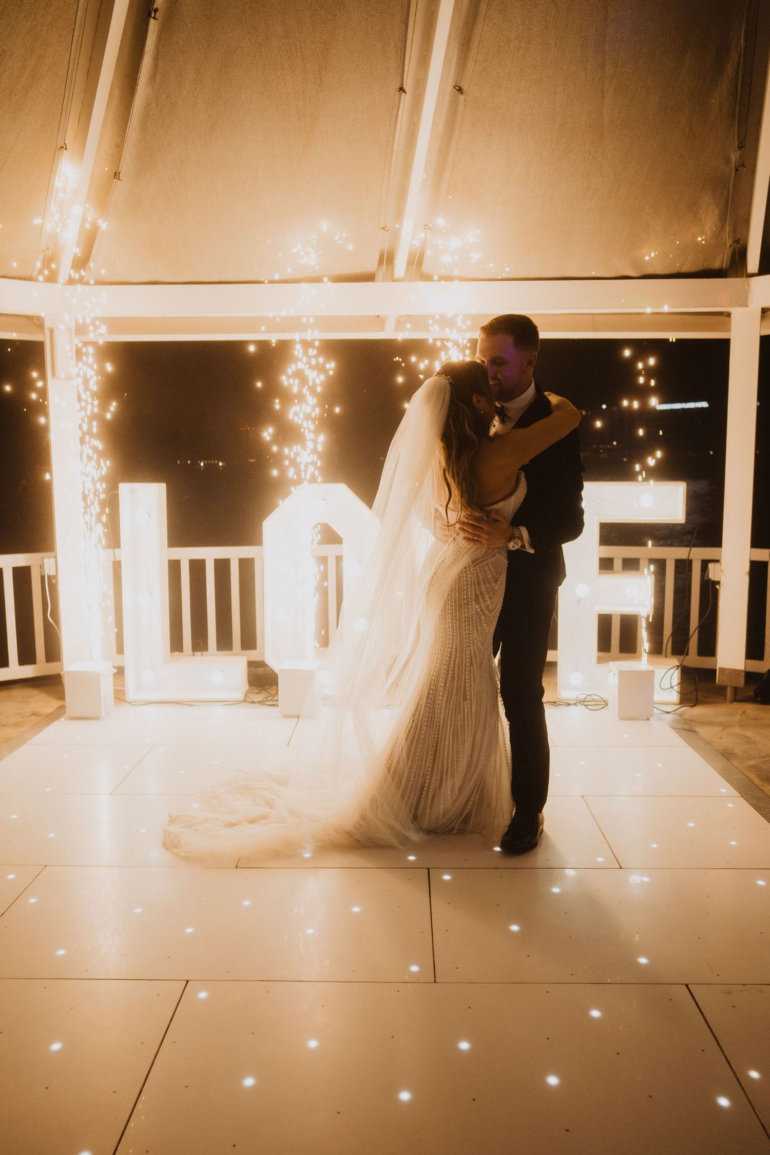 Bride and groom sharing their first dance on a lit dance floor with glowing LOVE letters and fairy lights.