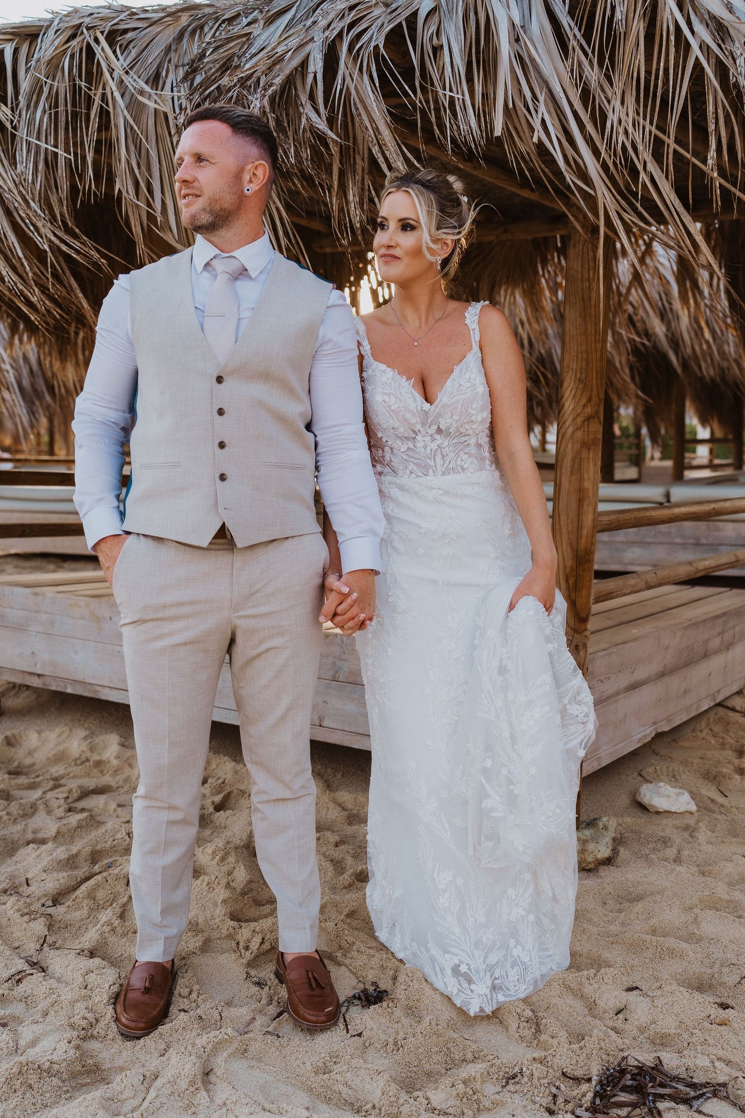 Newlyweds in beige and white attire pose together on a sandy beach under rustic thatched umbrellas.