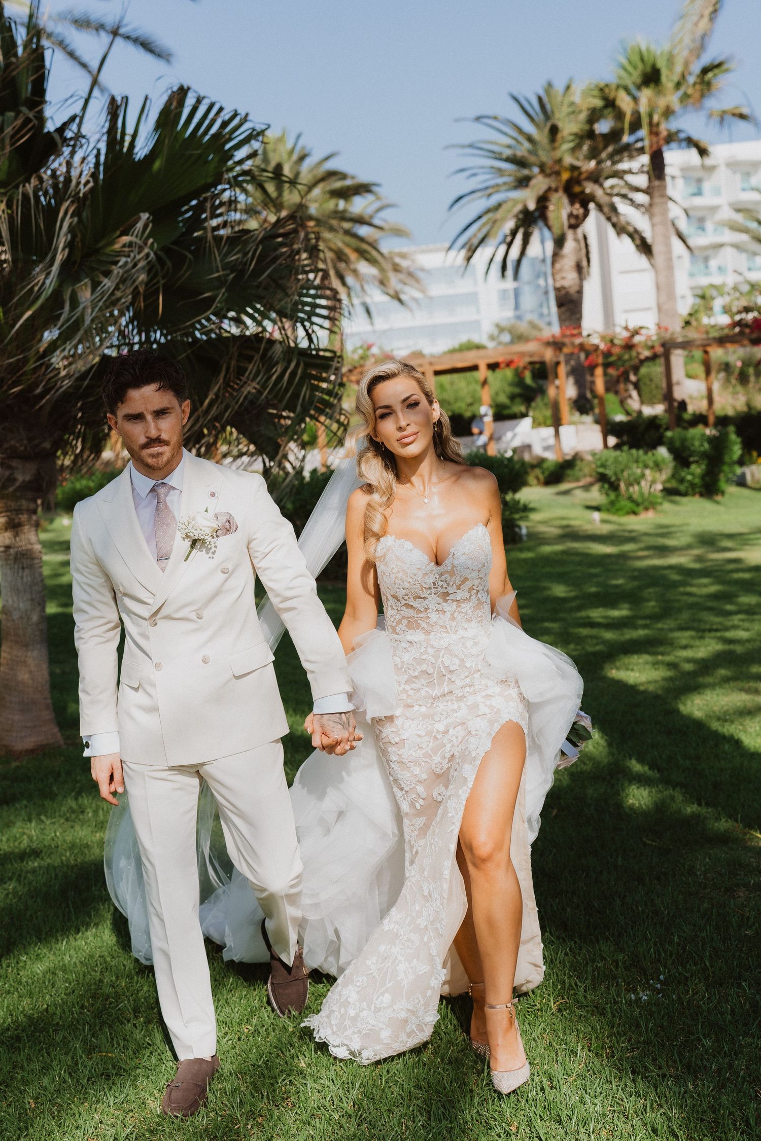 A couple walks on lush green lawn with palm trees, wearing a white suit and lace wedding dress with high slit.