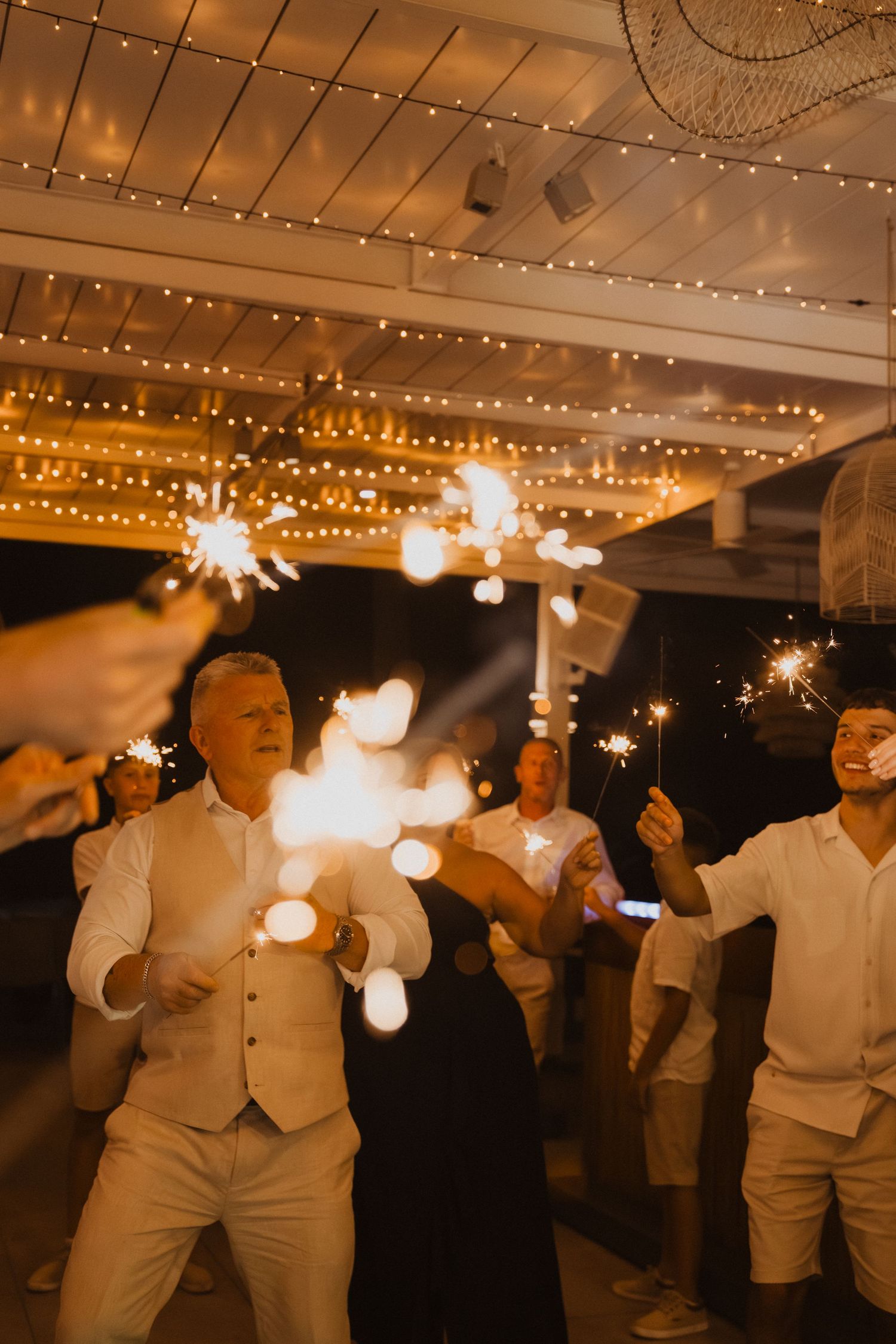 Wedding guests celebrate with sparklers under twinkling lights at an indoor reception venue.