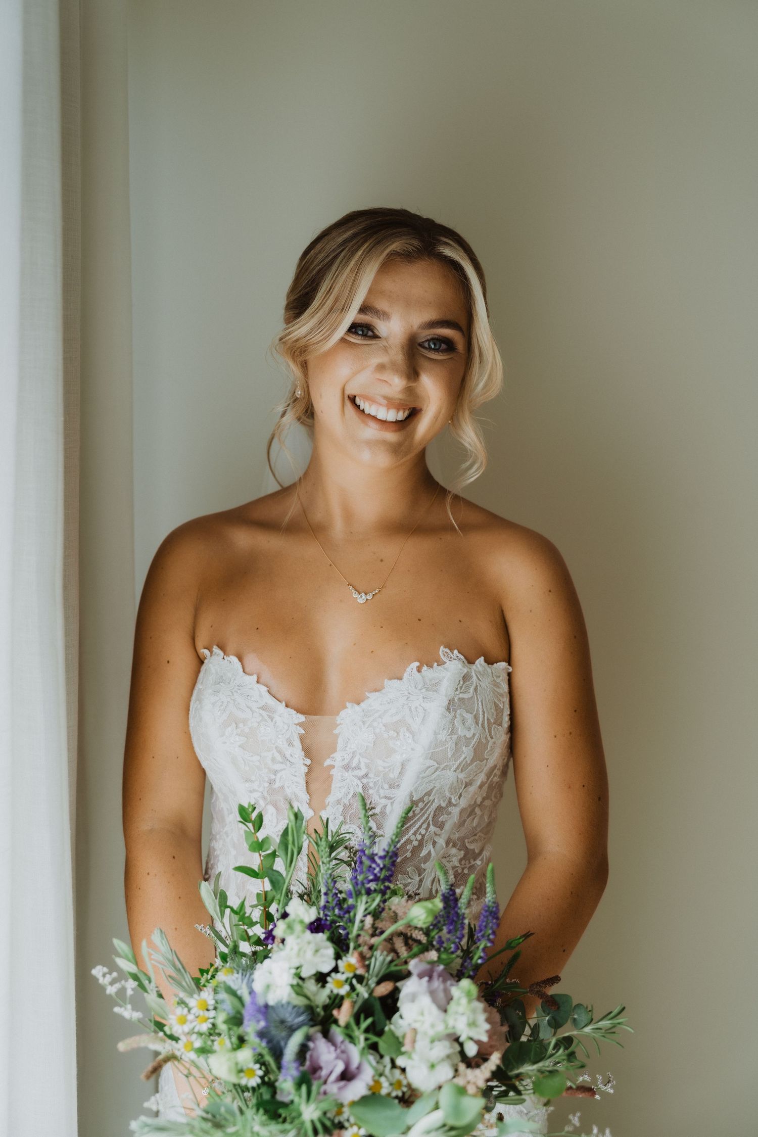Bride holding a fresh bouquet of purple, white and green flowers against a neutral background.