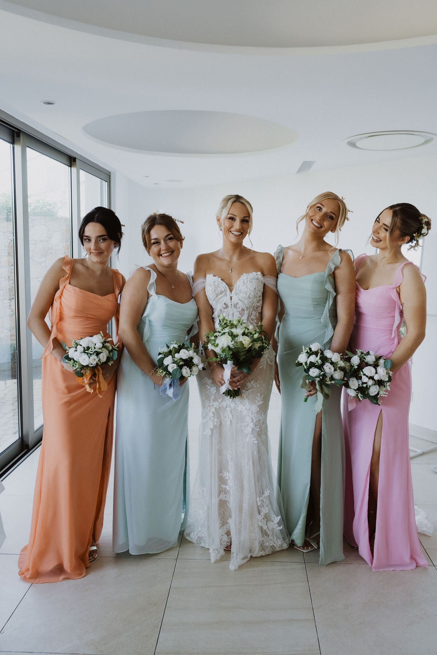 Wedding party in coral, mint, and pink dresses holding bouquets pose by large windows.