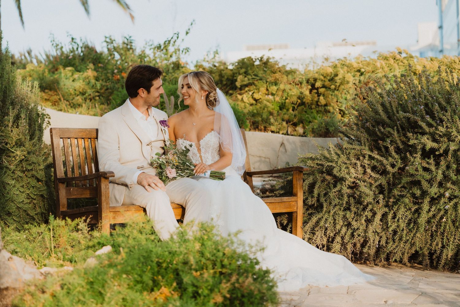 Newlyweds share a tender moment on a wooden bench surrounded by lush greenery and palm trees at sunset.