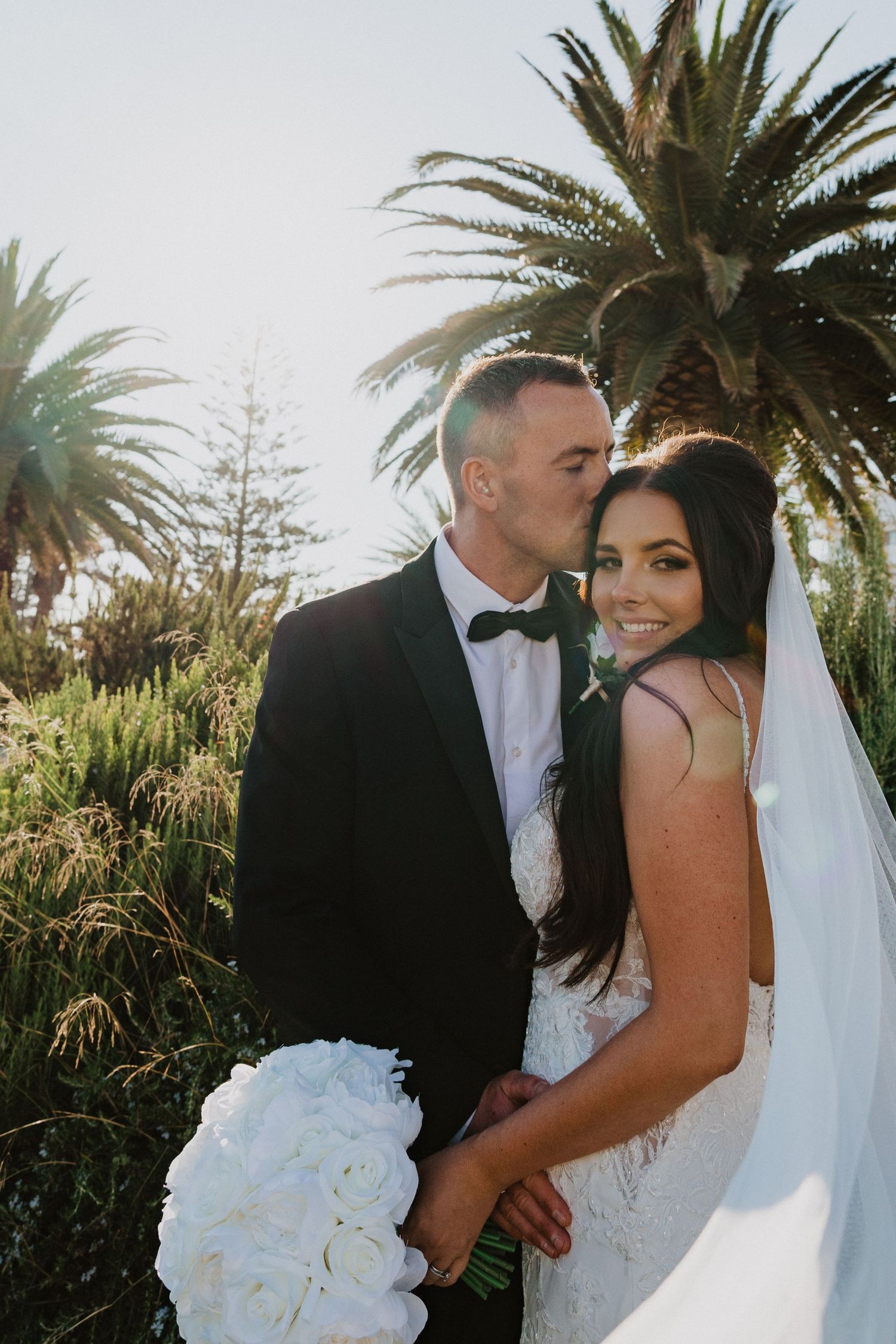 A wedding couple shares an intimate moment in a tropical garden setting with palm trees.