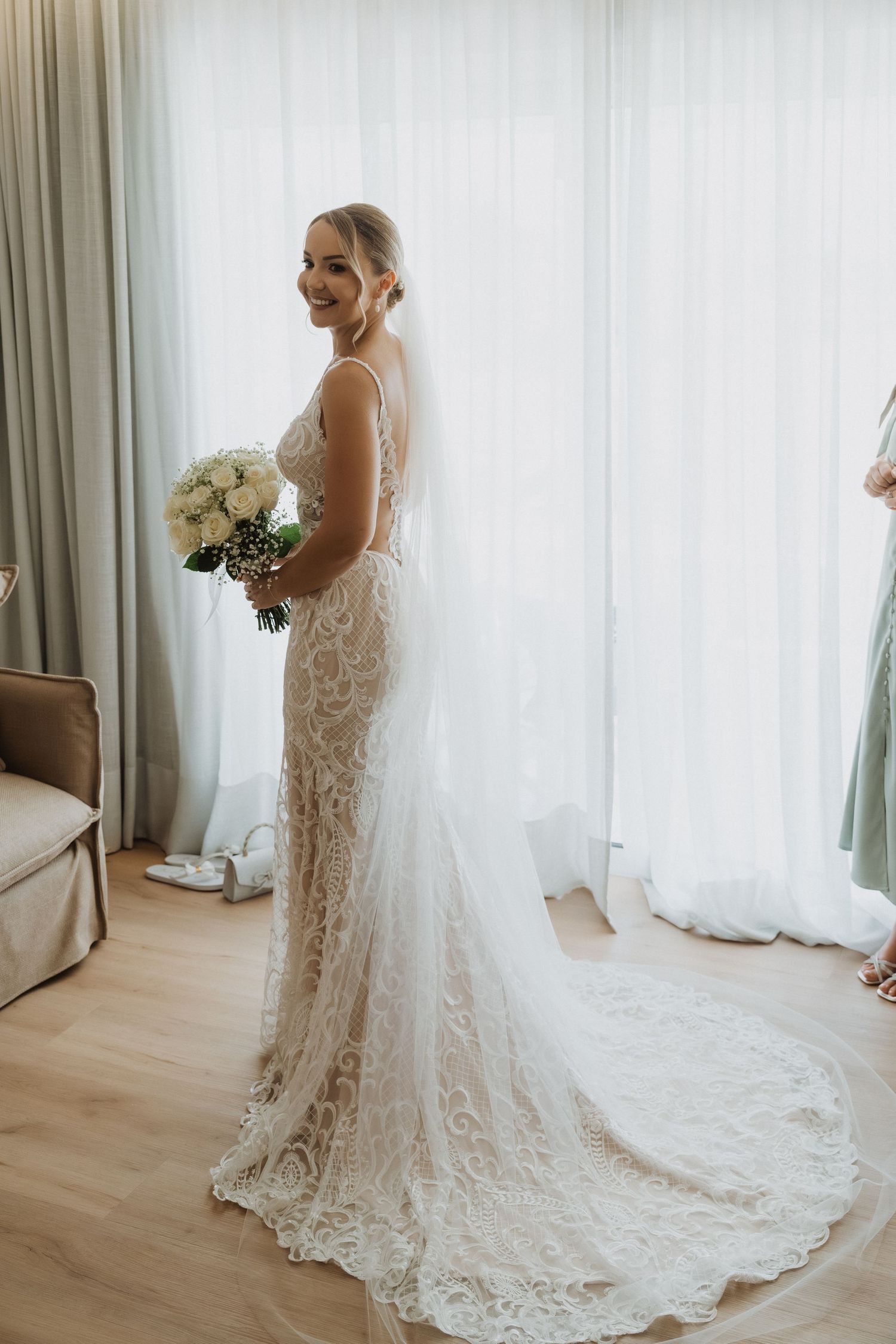 A stunning lace wedding gown with a flowing train is showcased in a bright room with sheer curtains.