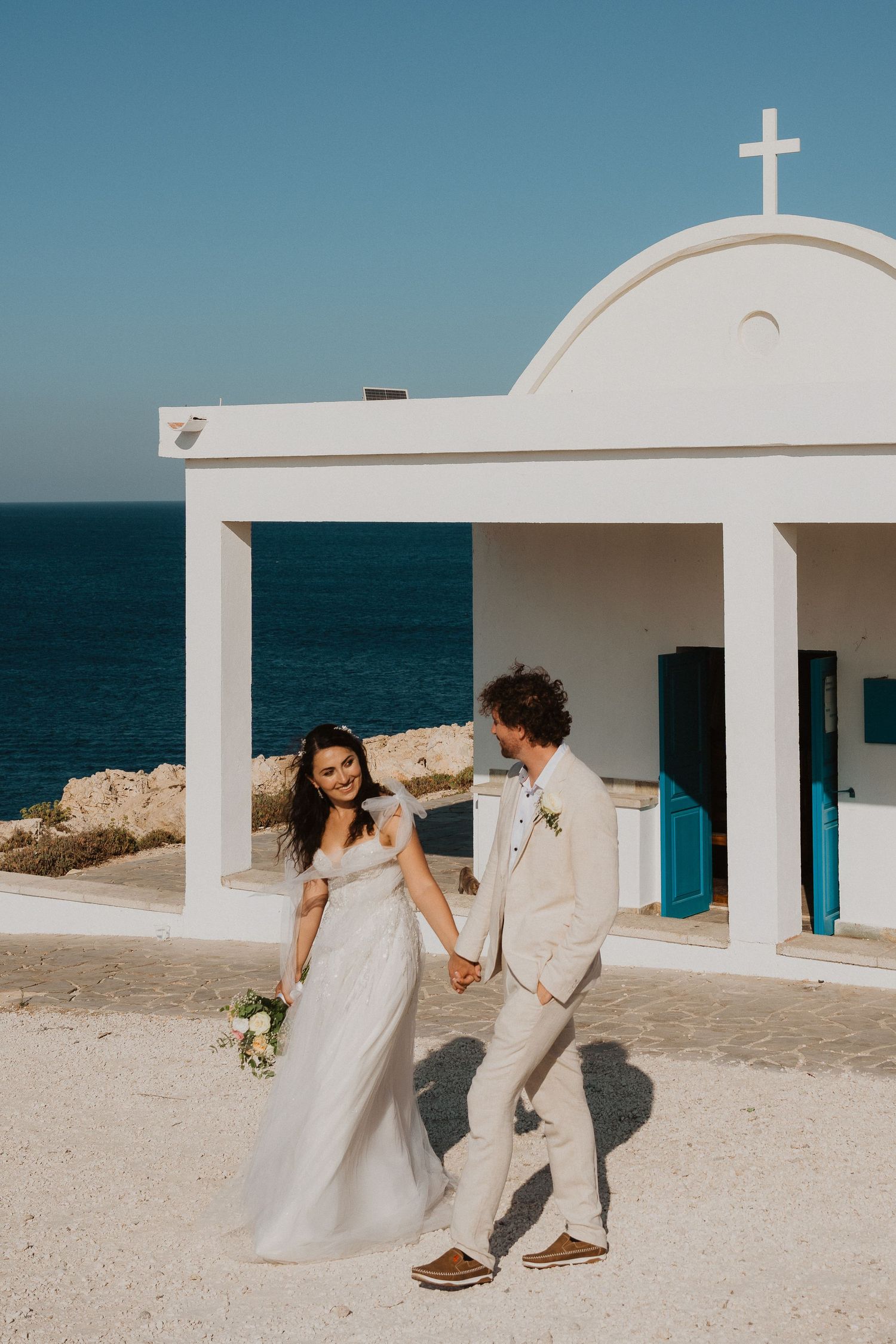 Couple in wedding attire standing by white Mediterranean chapel with ocean view.