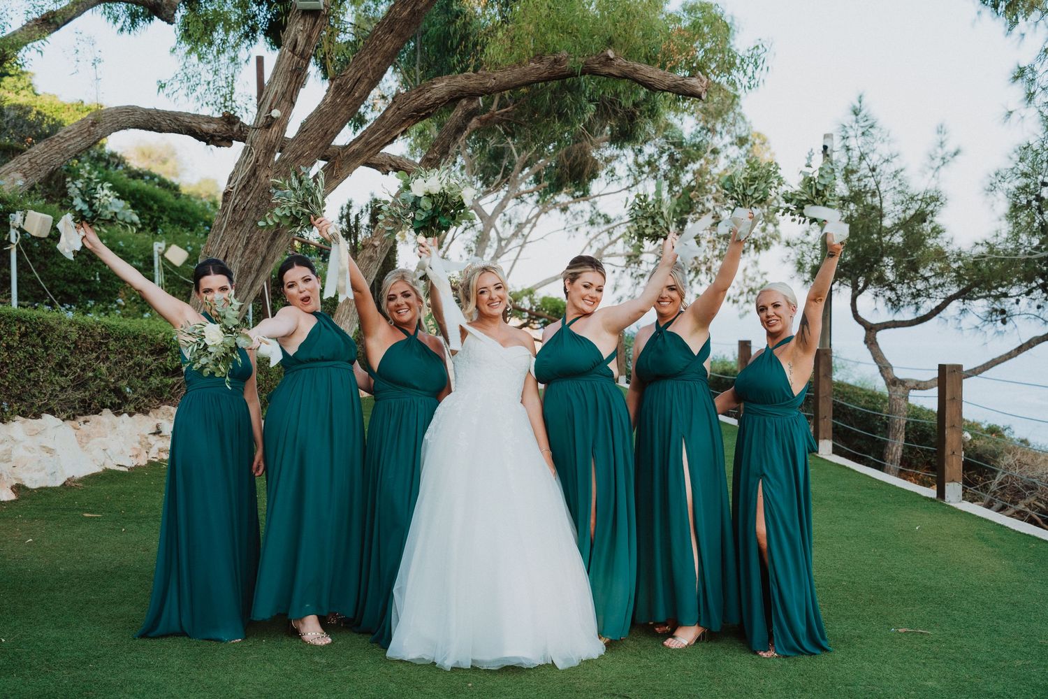 Wedding party in teal bridesmaid dresses celebrating joyfully on a grassy lawn under a large tree.