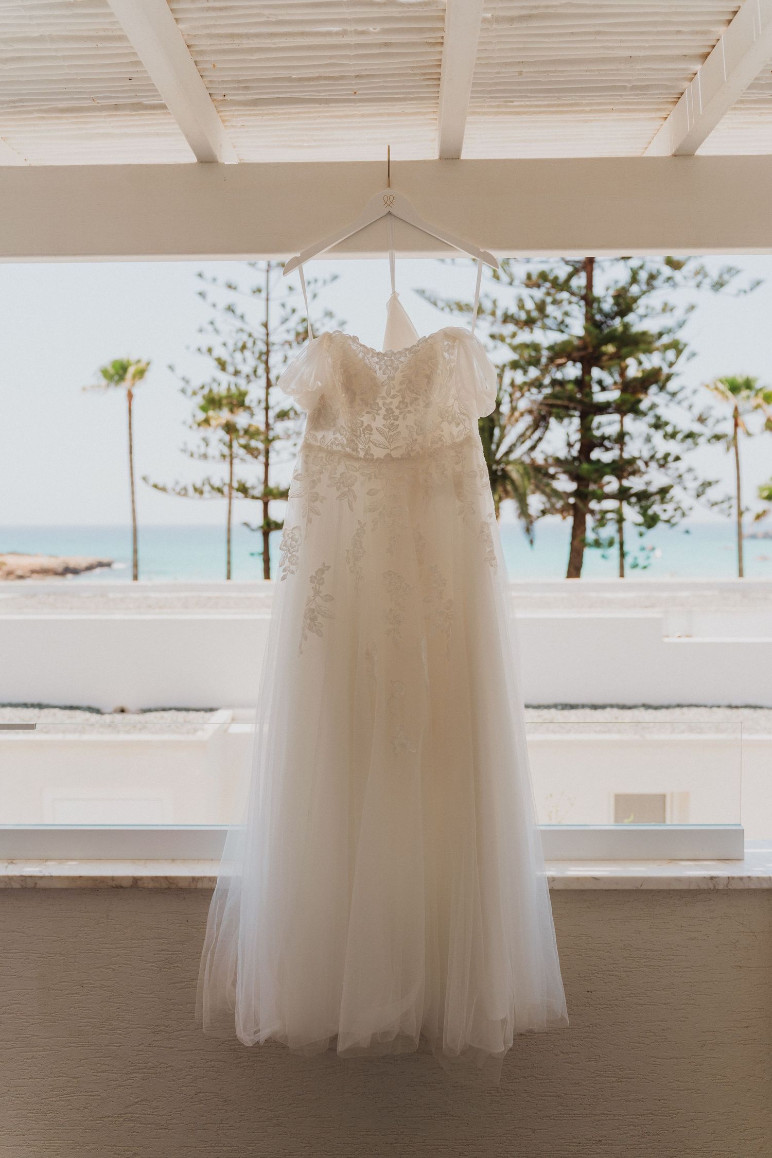 Beautiful wedding gown displayed on balcony overlooking ocean and palm trees.