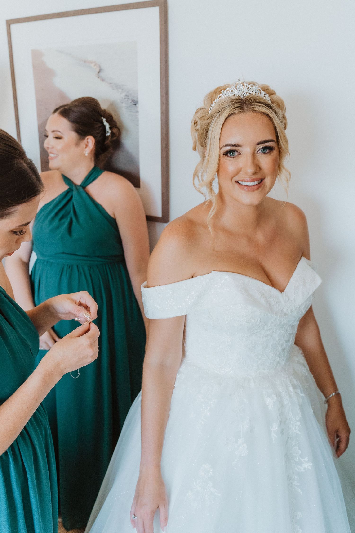 A bride wearing an off-shoulder white lace gown and crystal headpiece stands next to bridesmaid in emerald green halter dress.