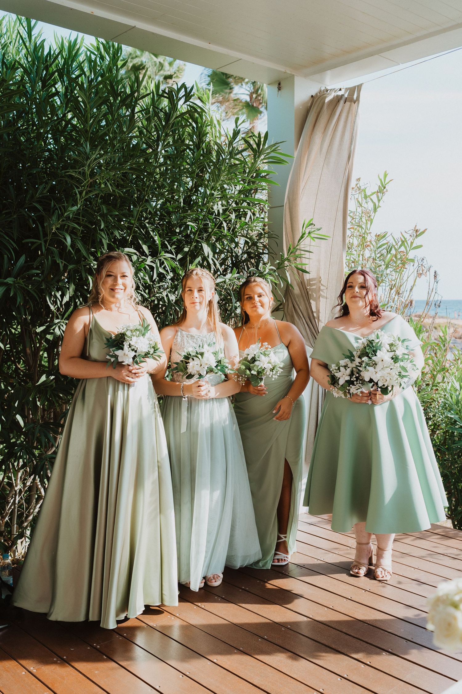 Bridesmaids in sage green and mint-colored dresses pose with white floral bouquets on a covered outdoor deck.