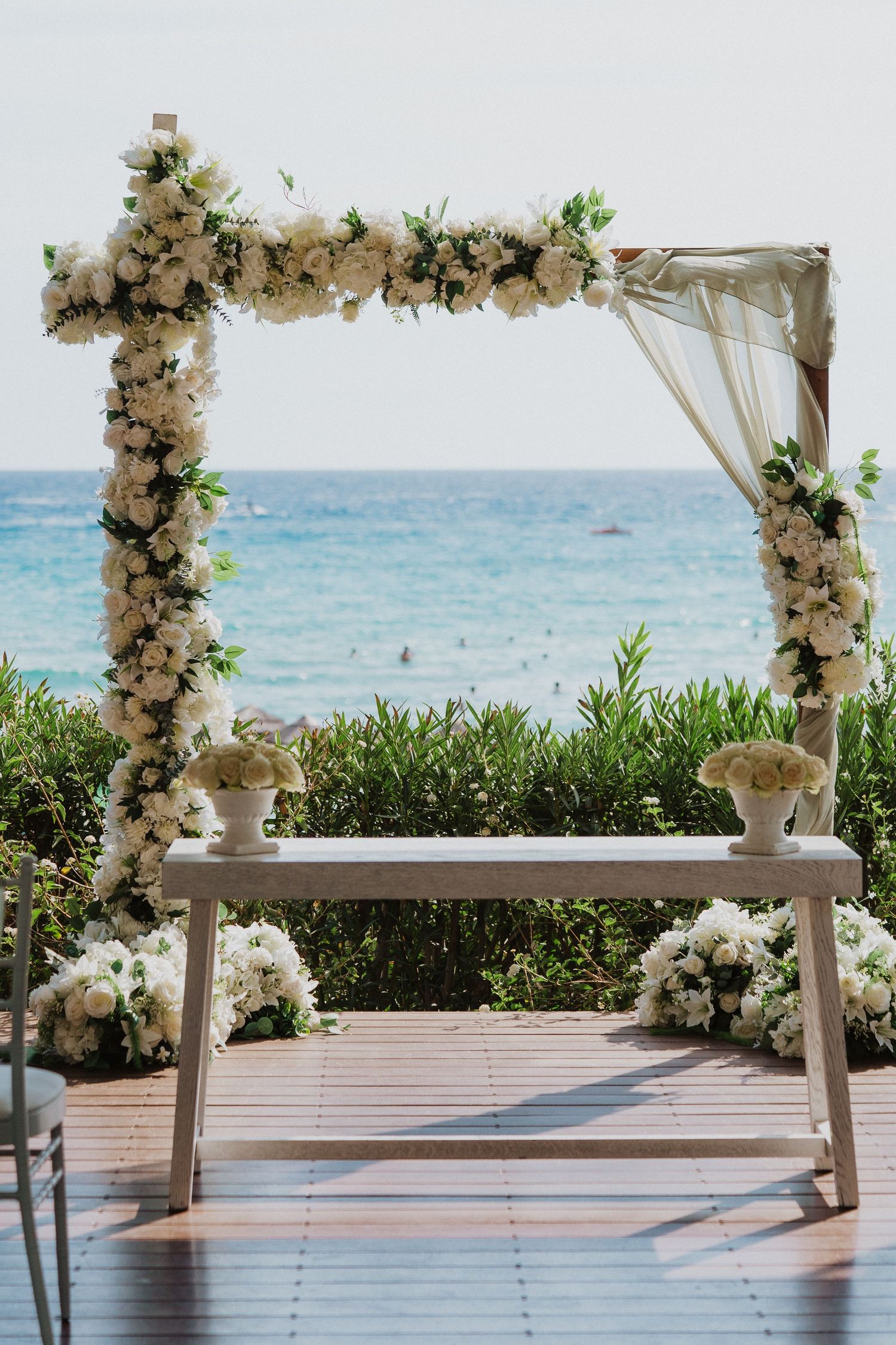 A romantic beachside wedding arch decorated with white flowers overlooks the ocean, with a wooden bench beneath it.
