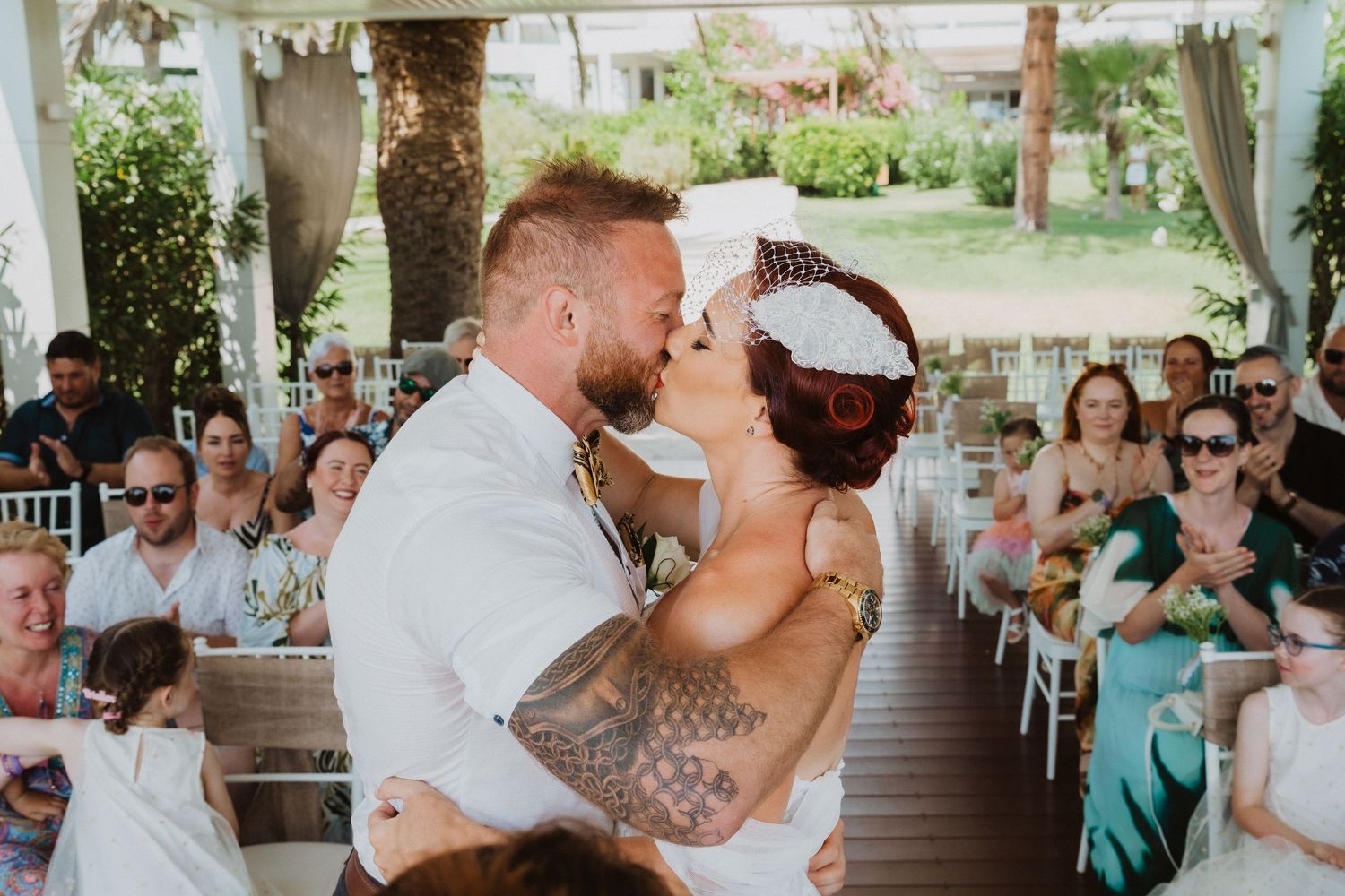 A couple shares their first kiss during an outdoor wedding ceremony surrounded by seated guests.