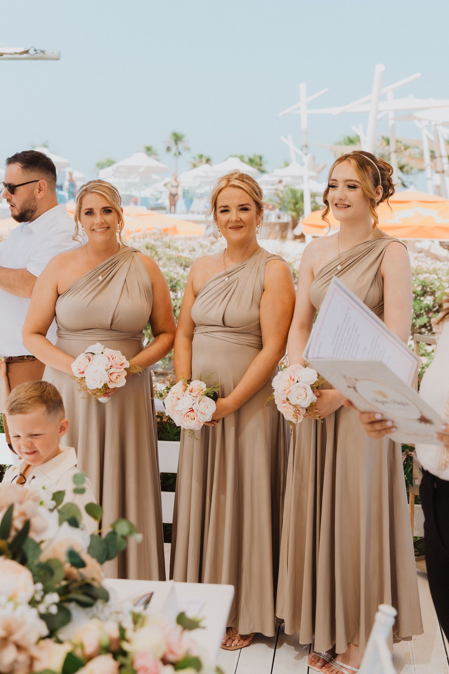 Bridesmaids in matching beige one-shoulder dresses holding white bouquets at outdoor ceremony.