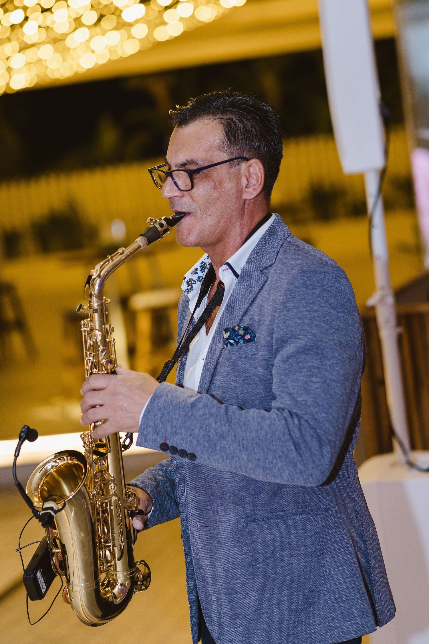 Saxophone player in grey blazer and glasses performs at an elegant event with bokeh lights.