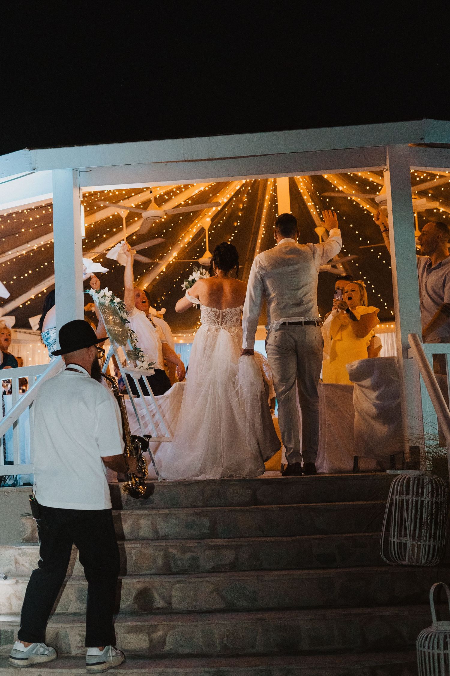 Wedding couple and guests celebrate under a gazebo decorated with warm glowing string lights at dusk.