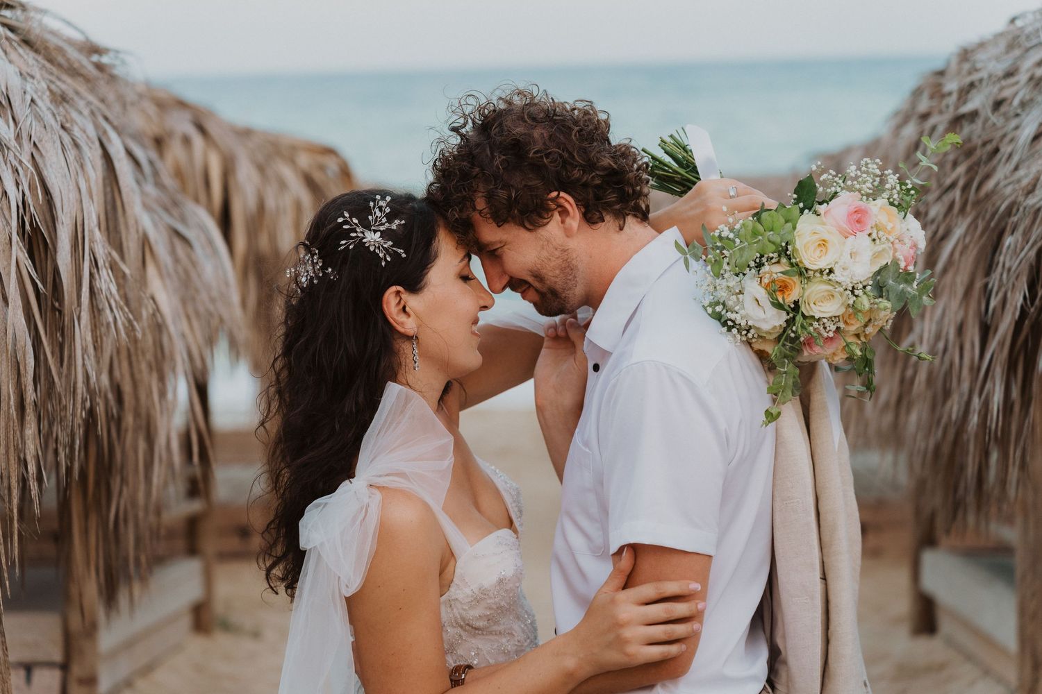 Intimate wedding portrait on beach boardwalk with thatched umbrellas and bouquet.