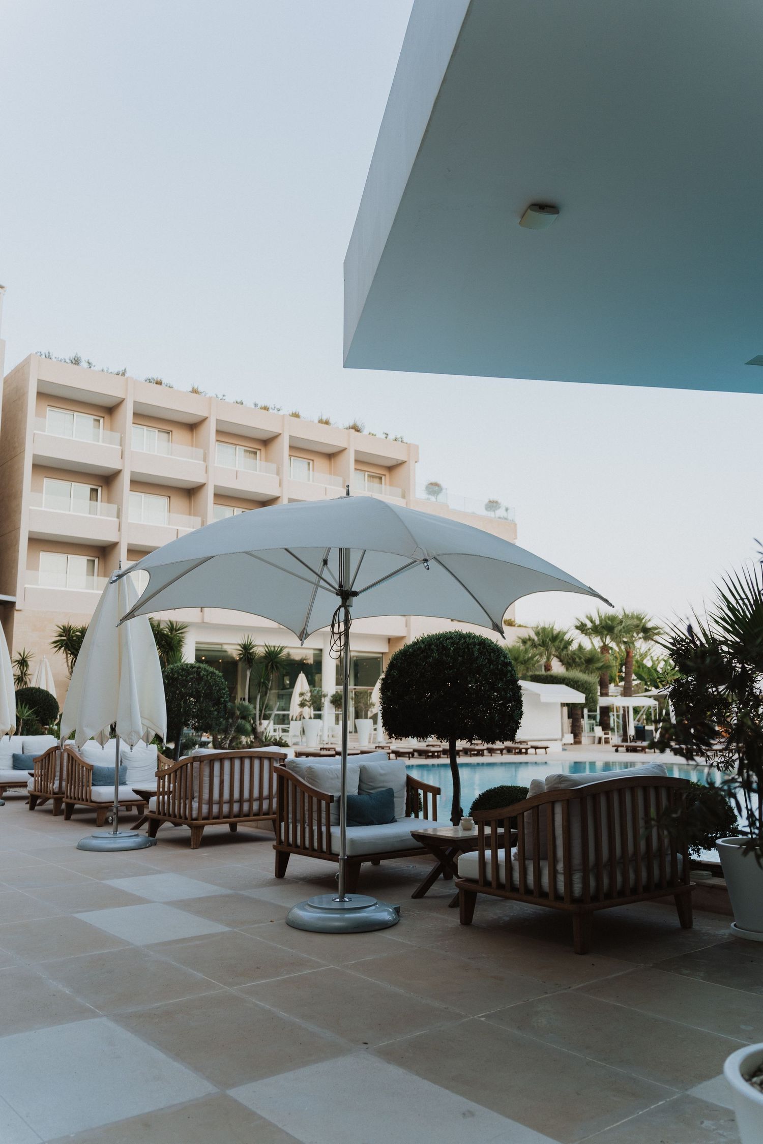 Modern hotel poolside lounge area with white umbrellas and wooden furniture on a tiled patio.