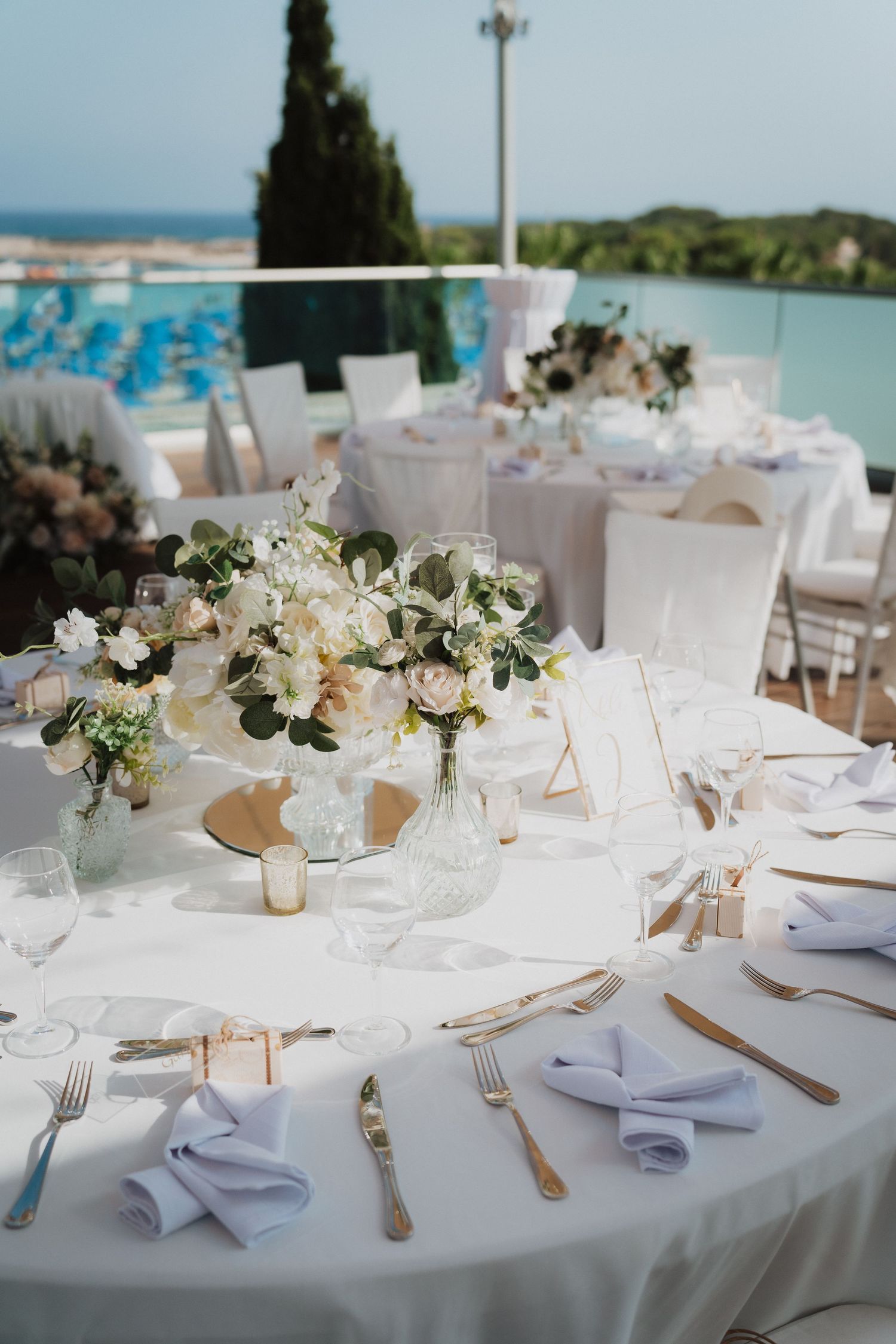 White and blush table setting with floral centerpieces and light blue napkins at waterfront venue.