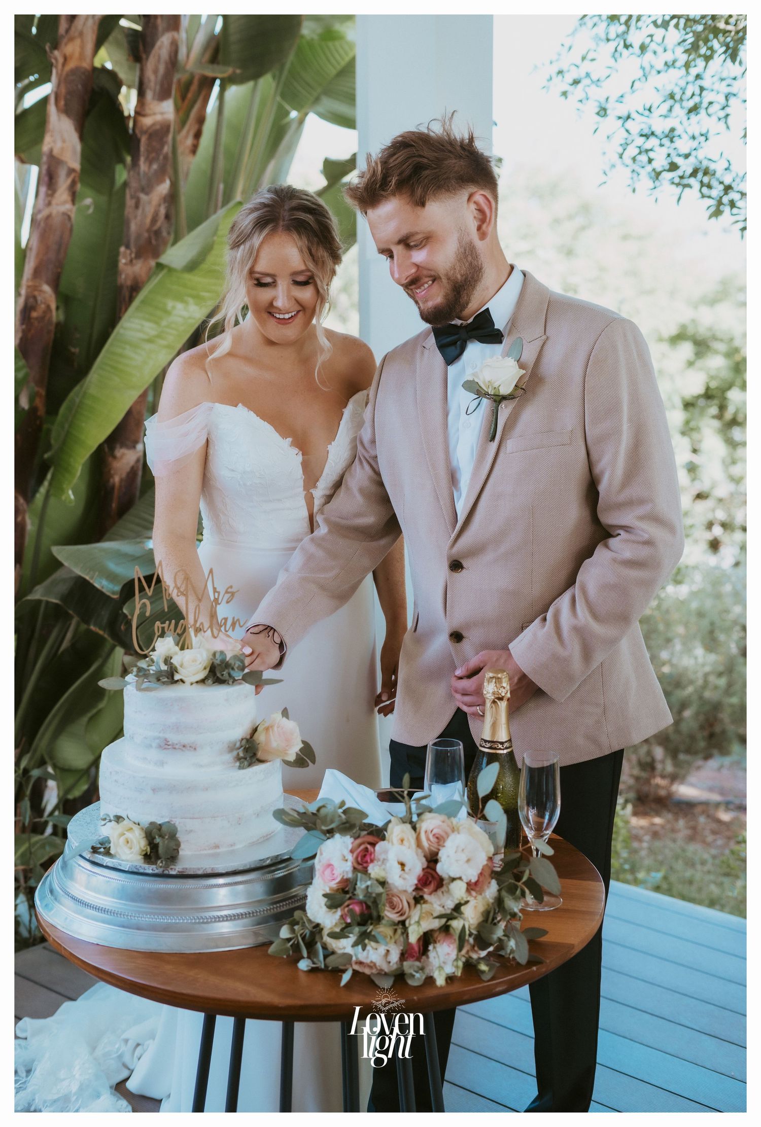 A couple cuts their elegant white wedding cake together with champagne and floral decorations on a wooden table.