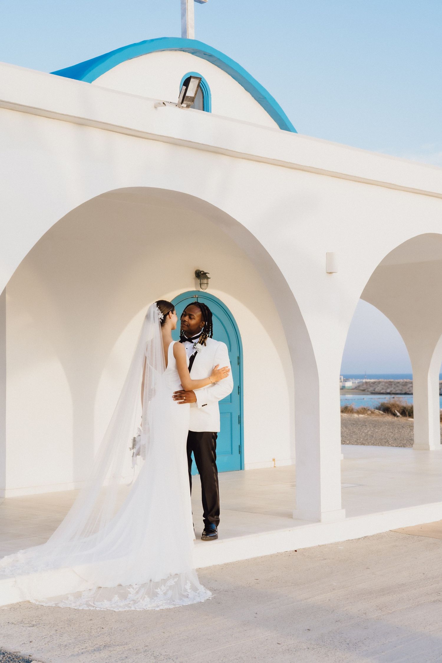 An intimate wedding portrait under white arches with a bright blue door and coastal backdrop.