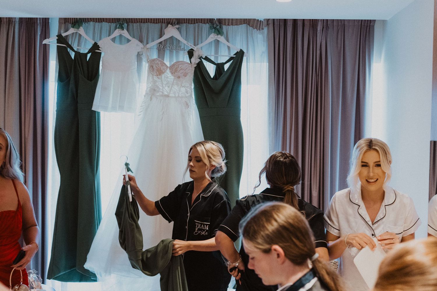 Multiple dark and light bridesmaid dresses hanging while people prepare for a wedding.