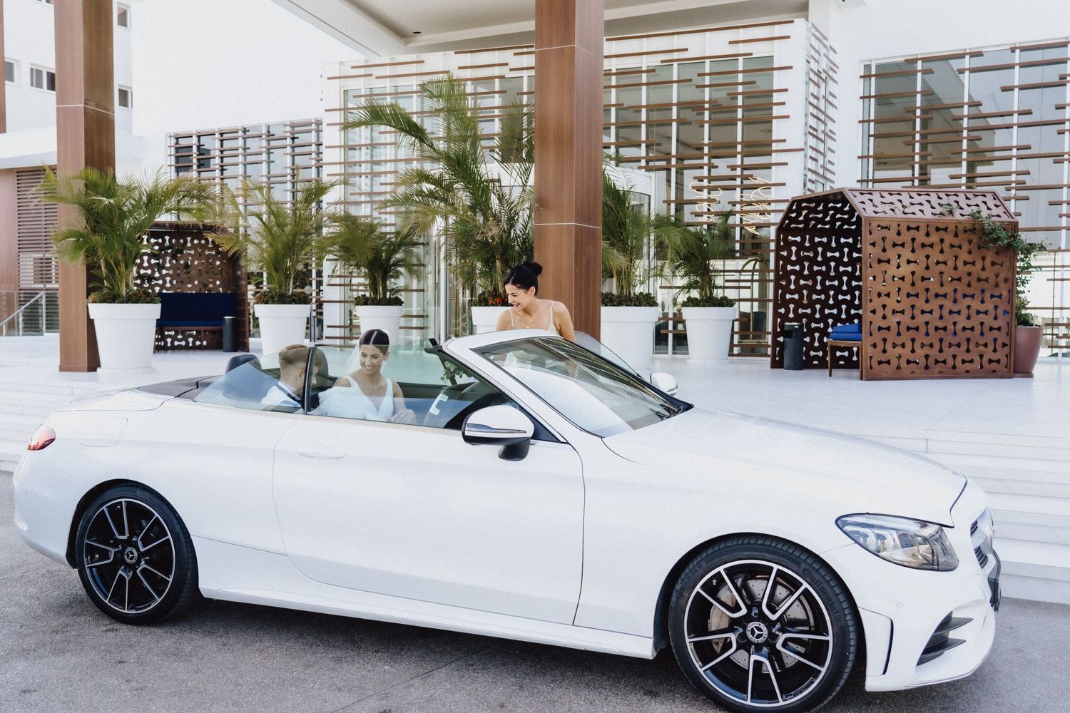 White Mercedes convertible parked in front of modern building with tropical palm decor.