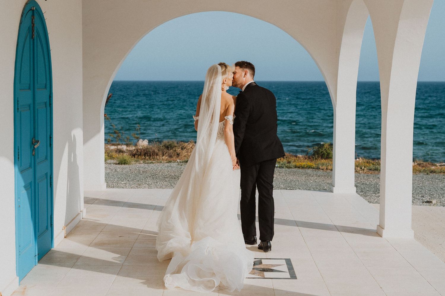 A seaside wedding portrait through Mediterranean-style white arches overlooking blue waters.