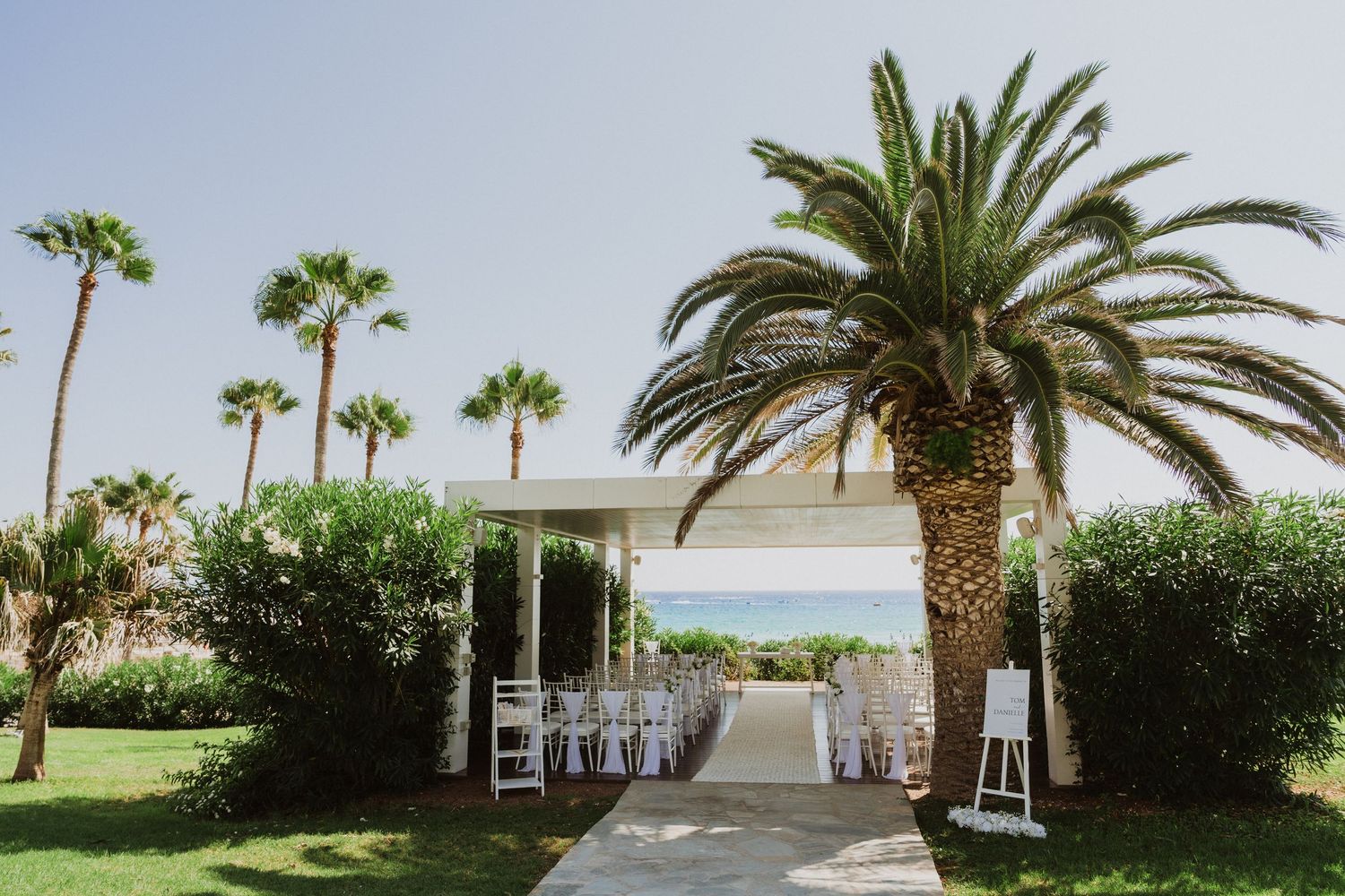 Palm trees line a white wedding ceremony walkway with white chairs and decorative lanterns.
