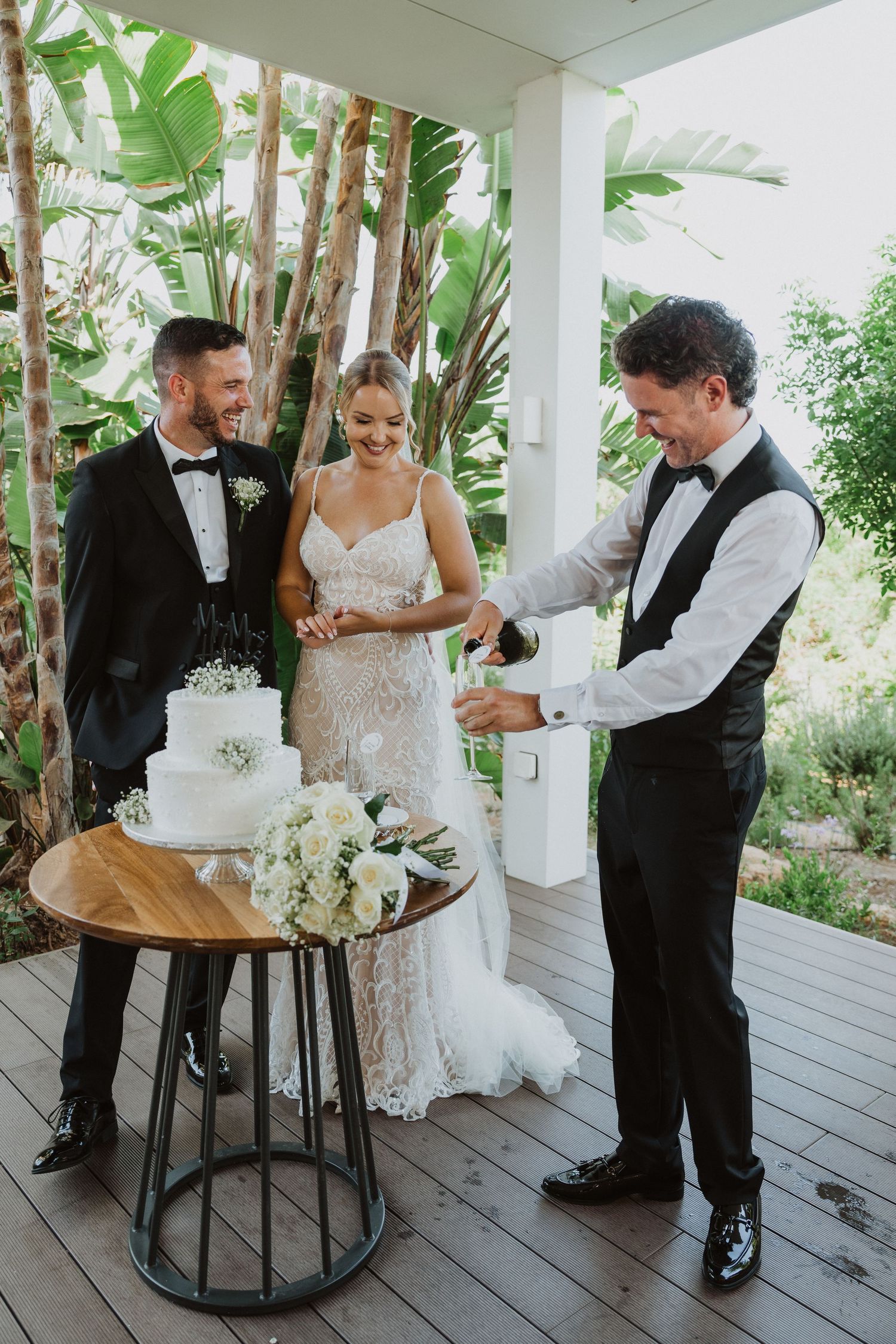 Newlyweds sharing a joyful cake cutting moment on a wooden deck surrounded by tropical plants.