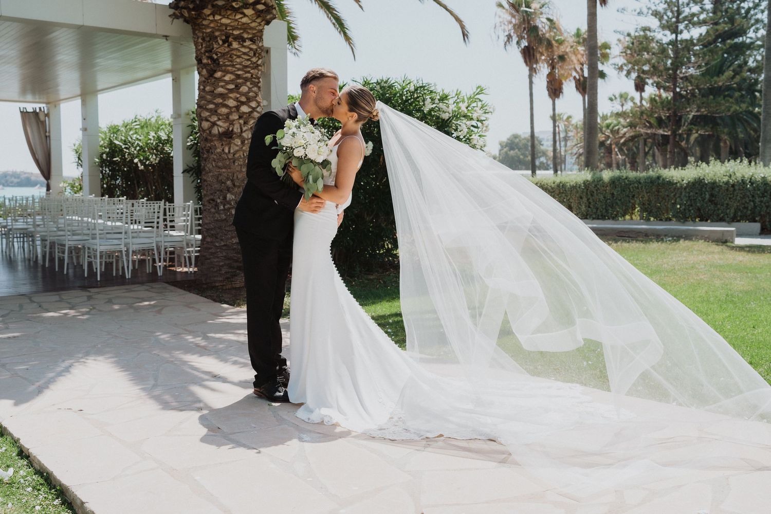 A wedding couple shares a romantic moment under a palm tree-lined walkway with a flowing white veil in the breeze.