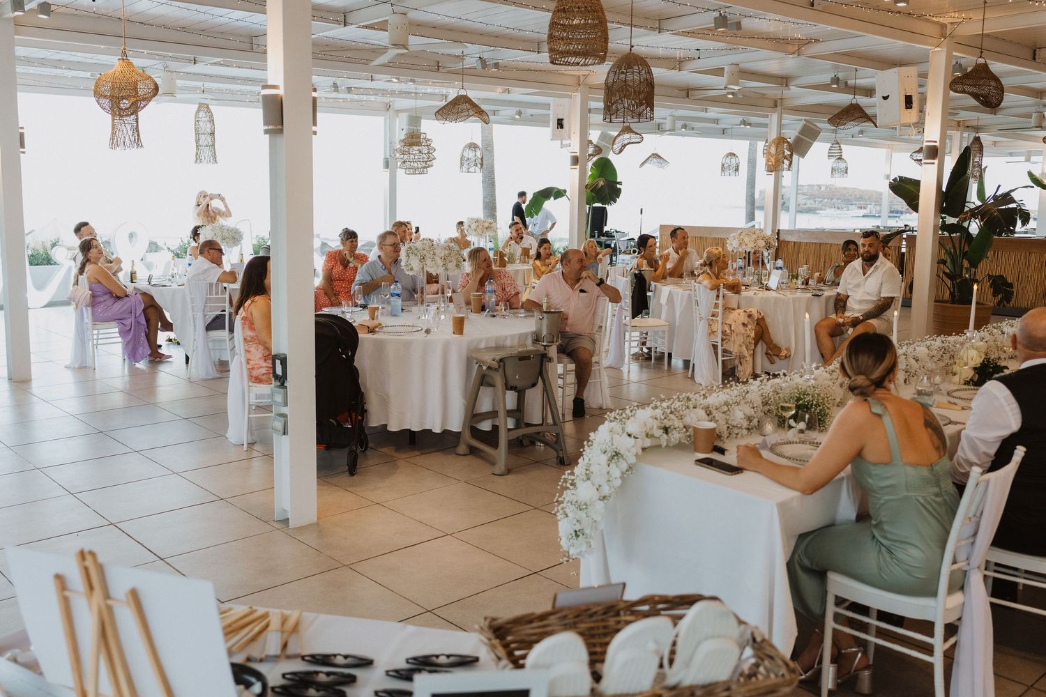 Bright, airy wedding reception venue with guests seated at decorated tables under rattan pendant lights.