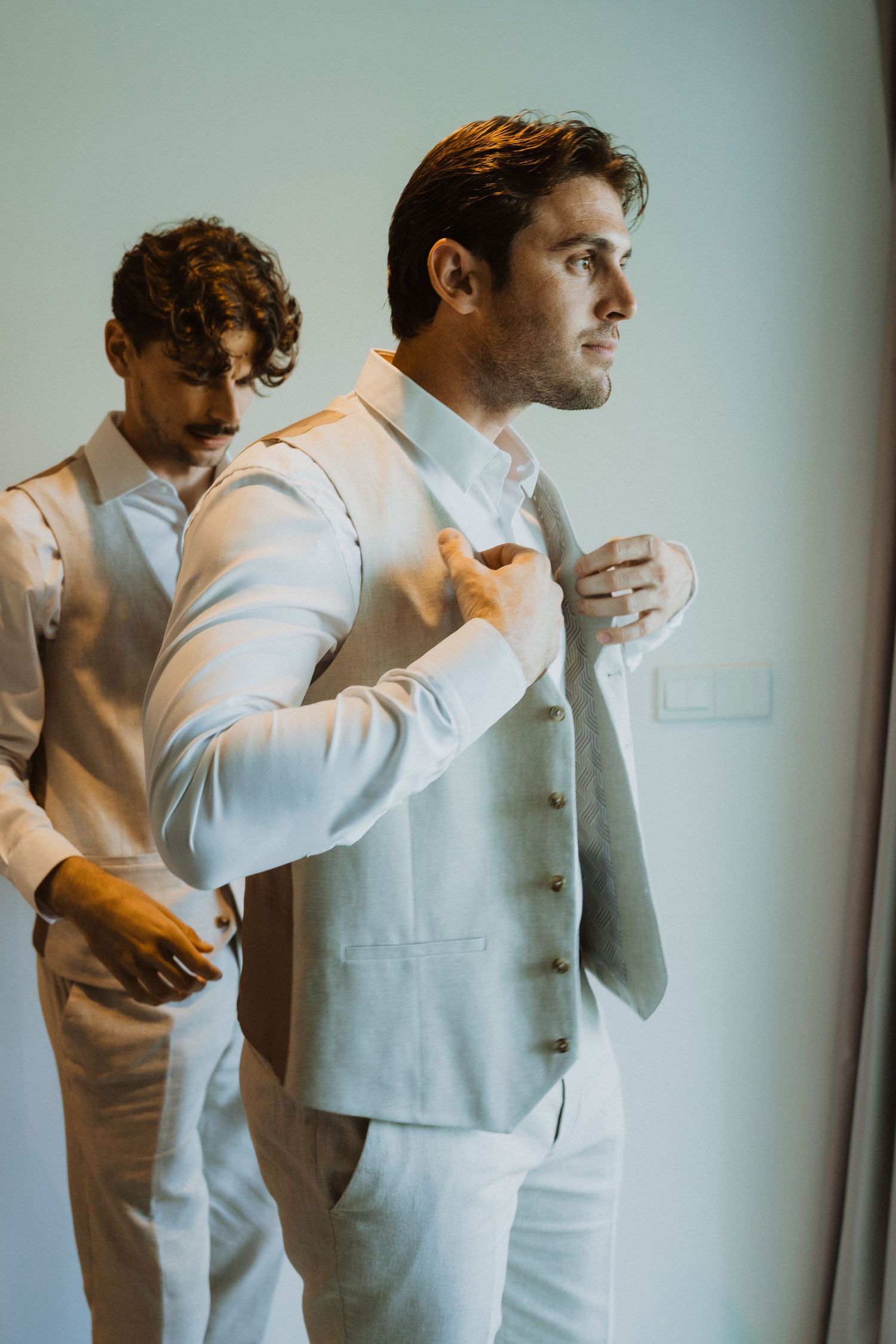 Two people in light colored formal suits adjusting their attire.
