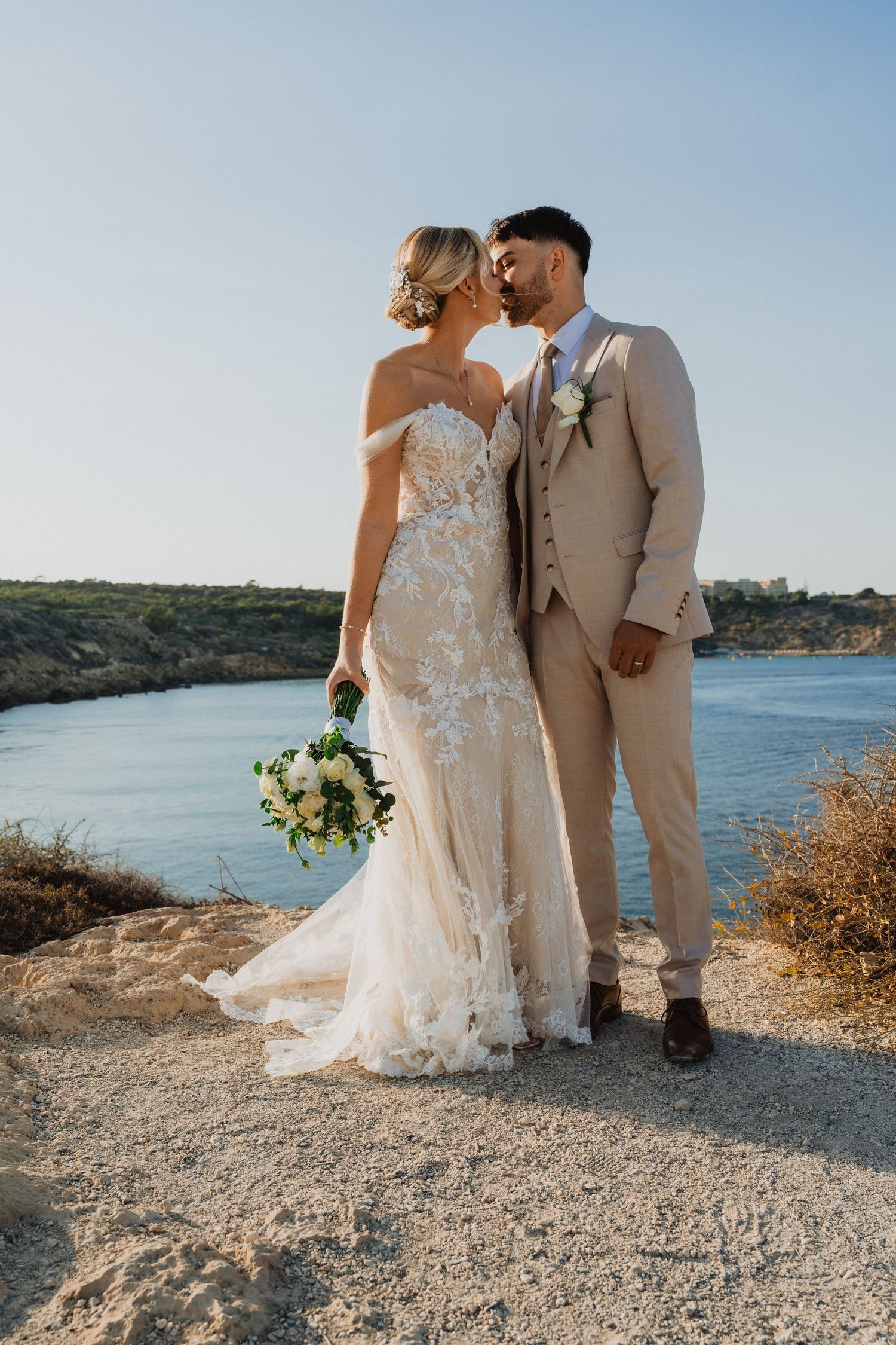 Newlyweds share an intimate moment on a seaside path with white wedding dress flowing in the breeze.
