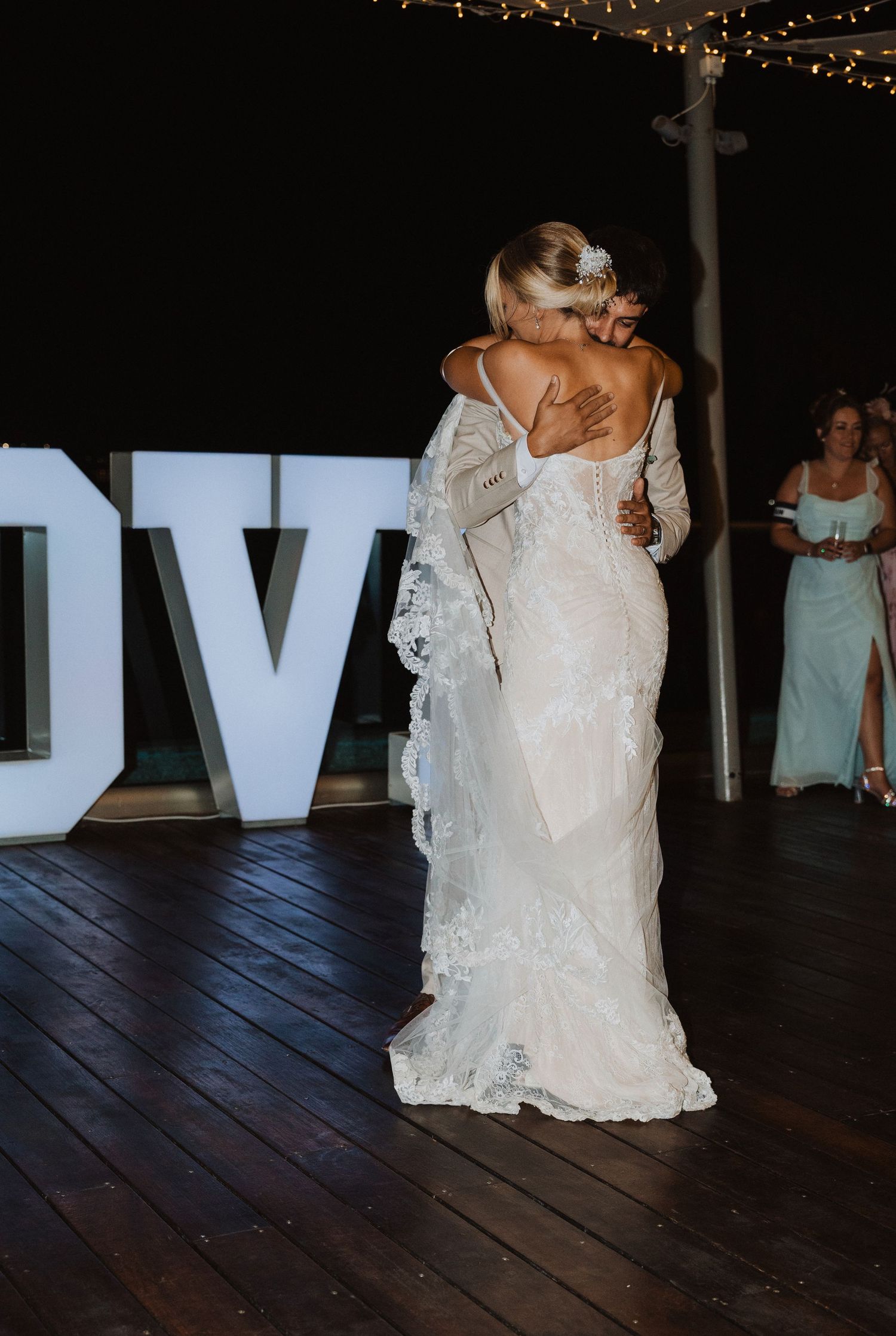 Bride in lace wedding gown poses by illuminated LOVE sign at evening reception.