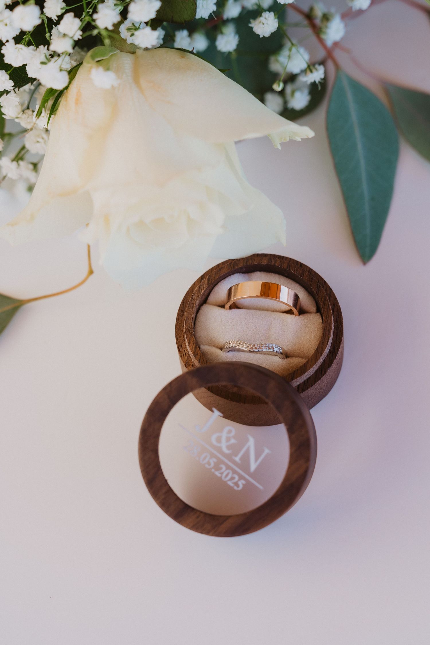 Wedding rings nestled in brown velvet ring boxes surrounded by white flowers and green leaves on a soft pink background.
