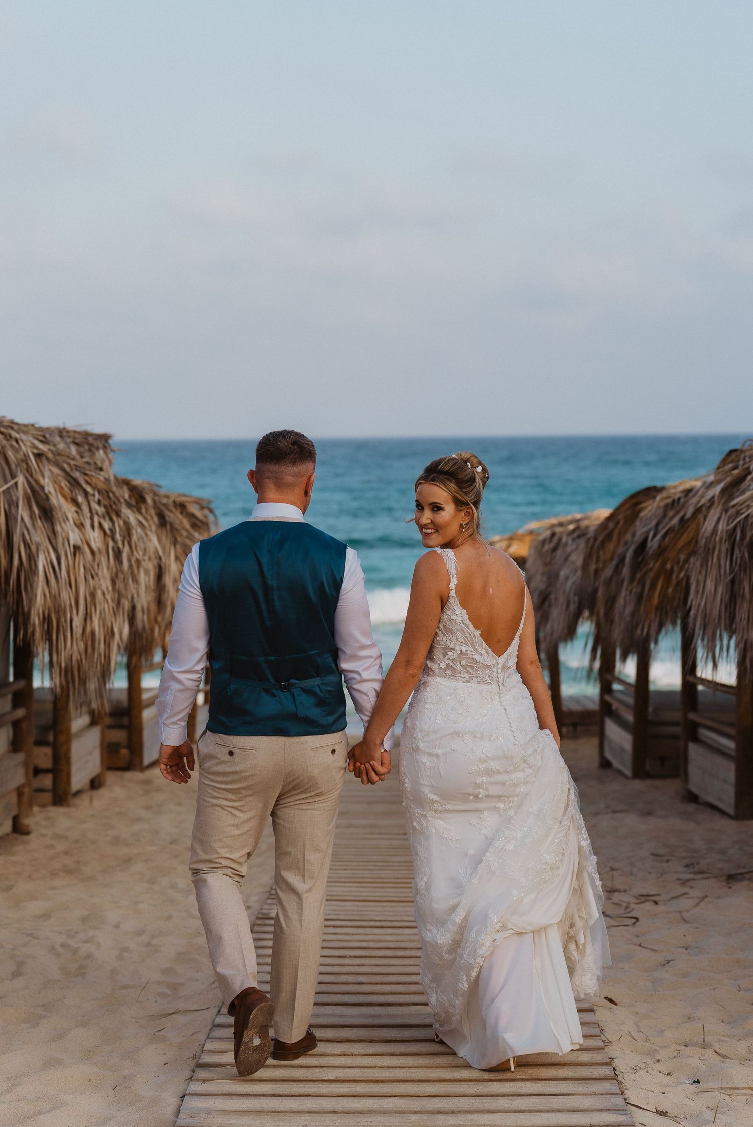 Couple walking hand in hand down a wooden boardwalk lined with palm umbrellas on a tropical beach at sunset.