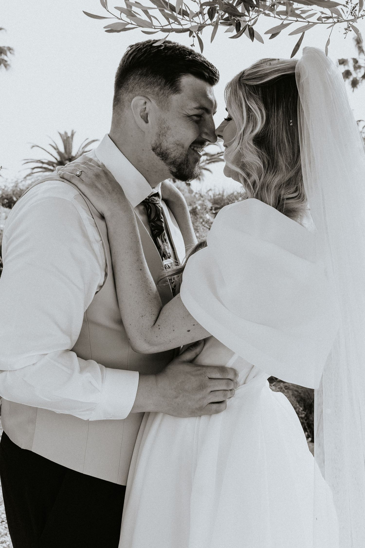 Intimate black and white portrait of wedding couple sharing tender moment under olive branches.