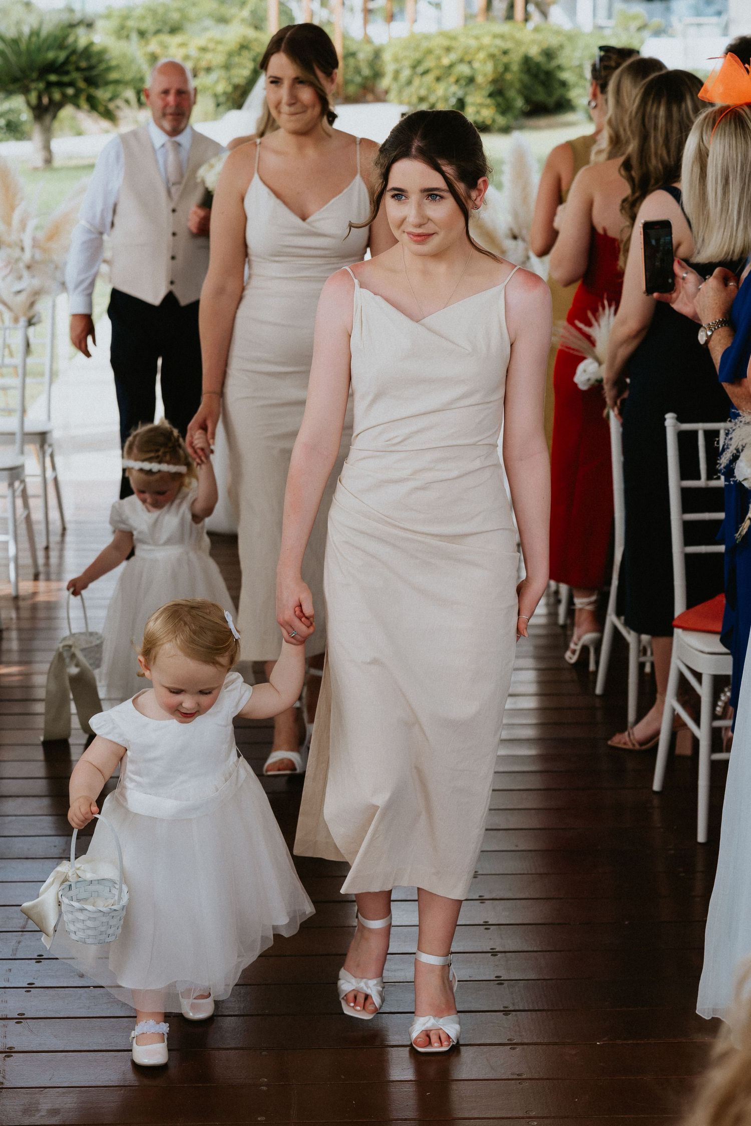 Bridesmaids in neutral dresses walk down wooden aisle with flower girls at outdoor ceremony.