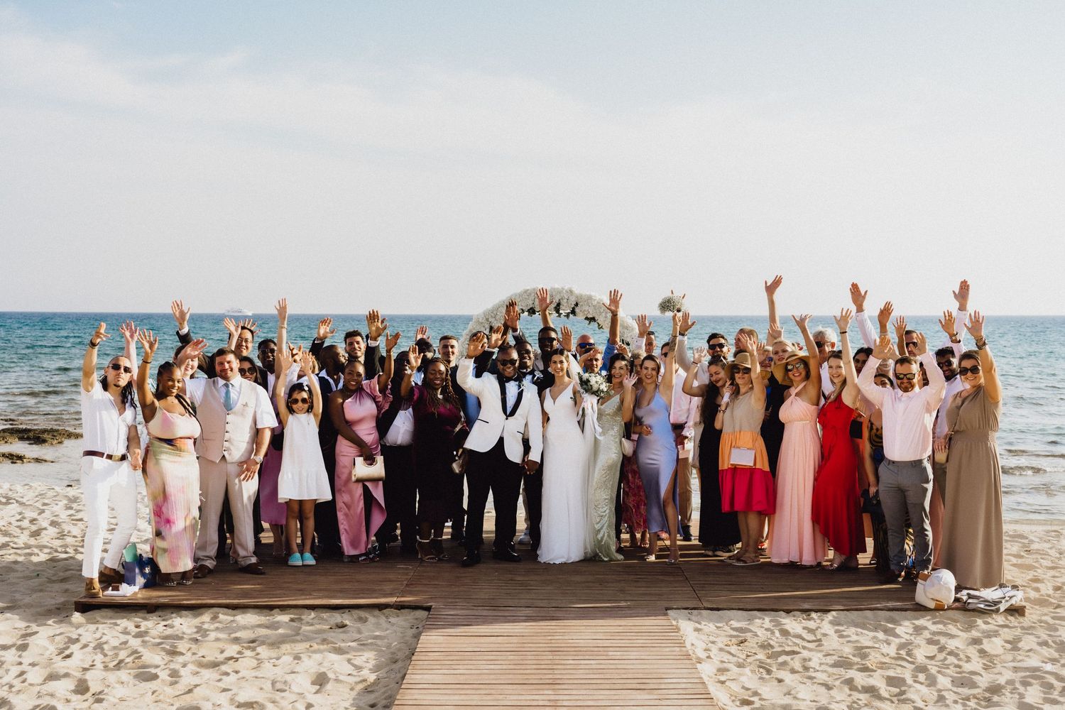 Large wedding party group celebrates together on a beach boardwalk at sunset.