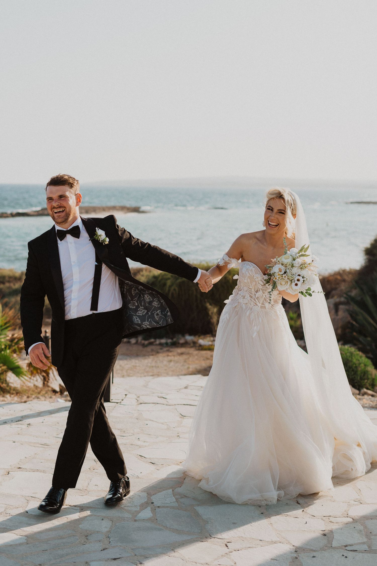 Wedding couple walks hand in hand along coastal path with ocean views.