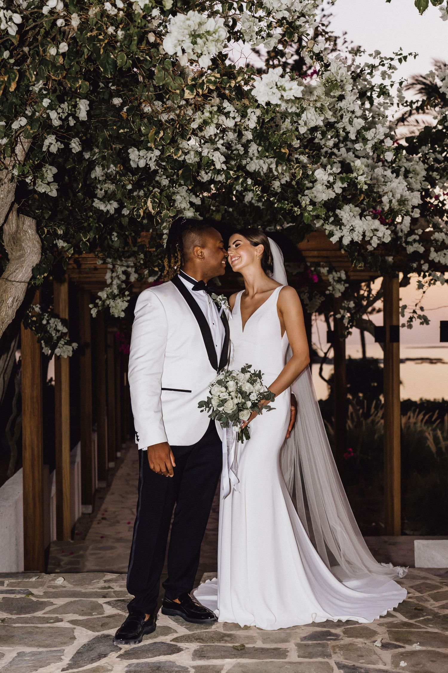 A wedding couple poses under blooming white flowers on a stone pathway during sunset.