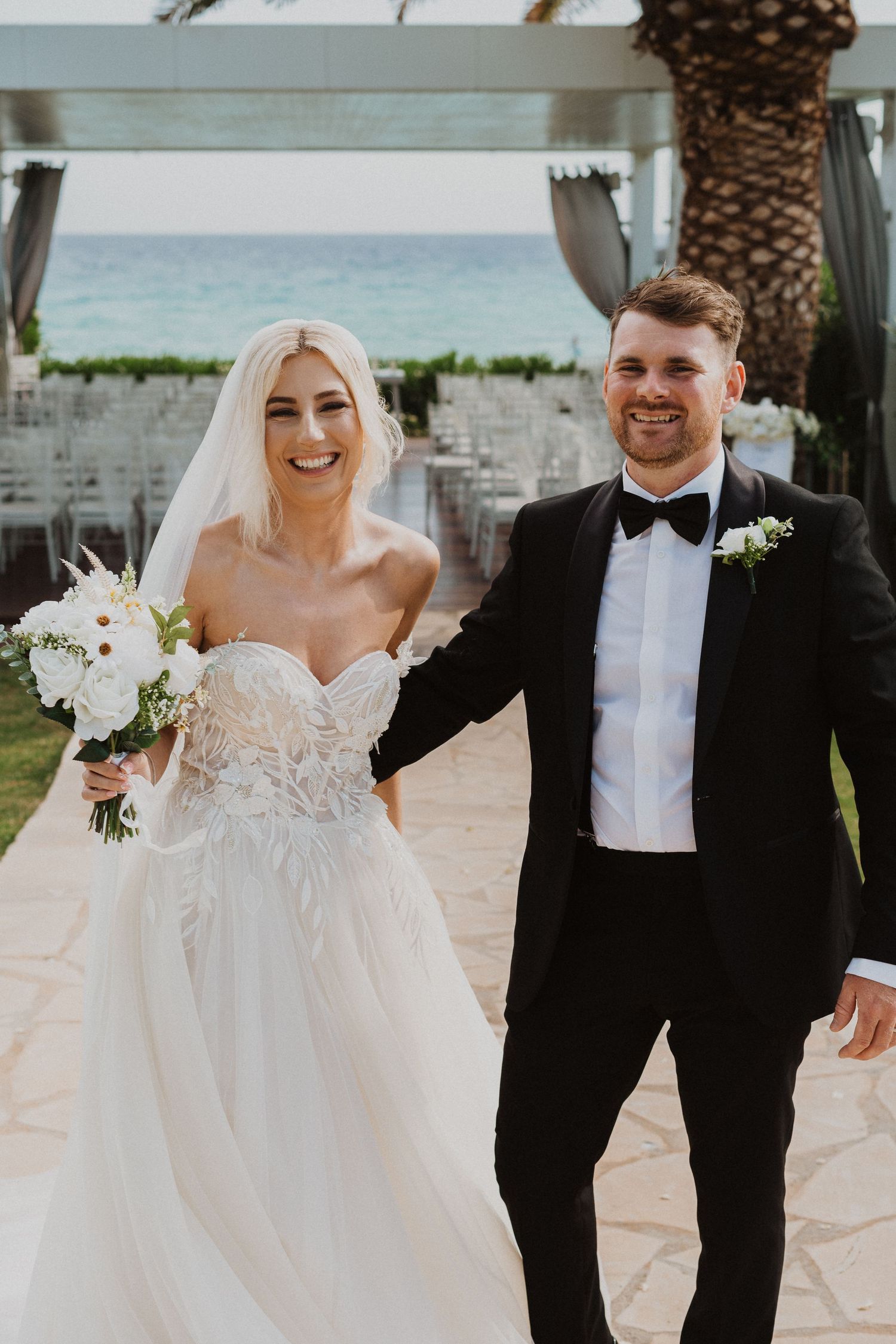 Joyful wedding couple celebrates under white pavilion with ocean and palm trees in background.